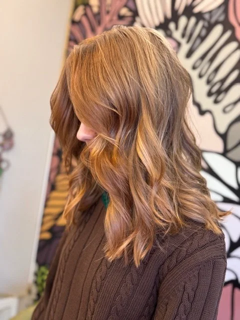 Side view of a woman with wavy, shoulder-length, light brown hair with highlights, wearing a brown knitted sweater, in front of a colorful abstract wall art background.