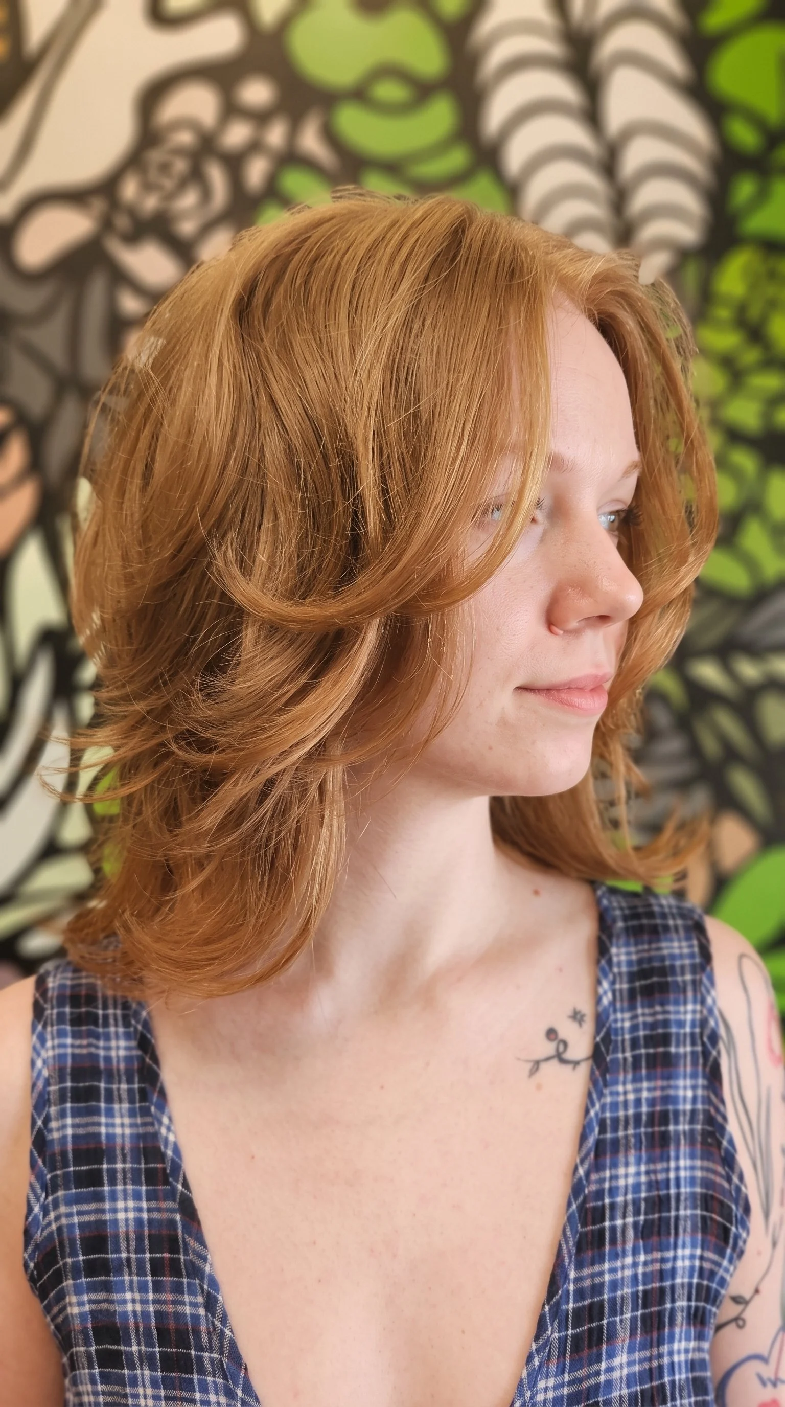 Close-up portrait of a person with shoulder-length copper-red hair wearing a blue plaid sleeveless top, with visible tattoos and a leafy graphic background.