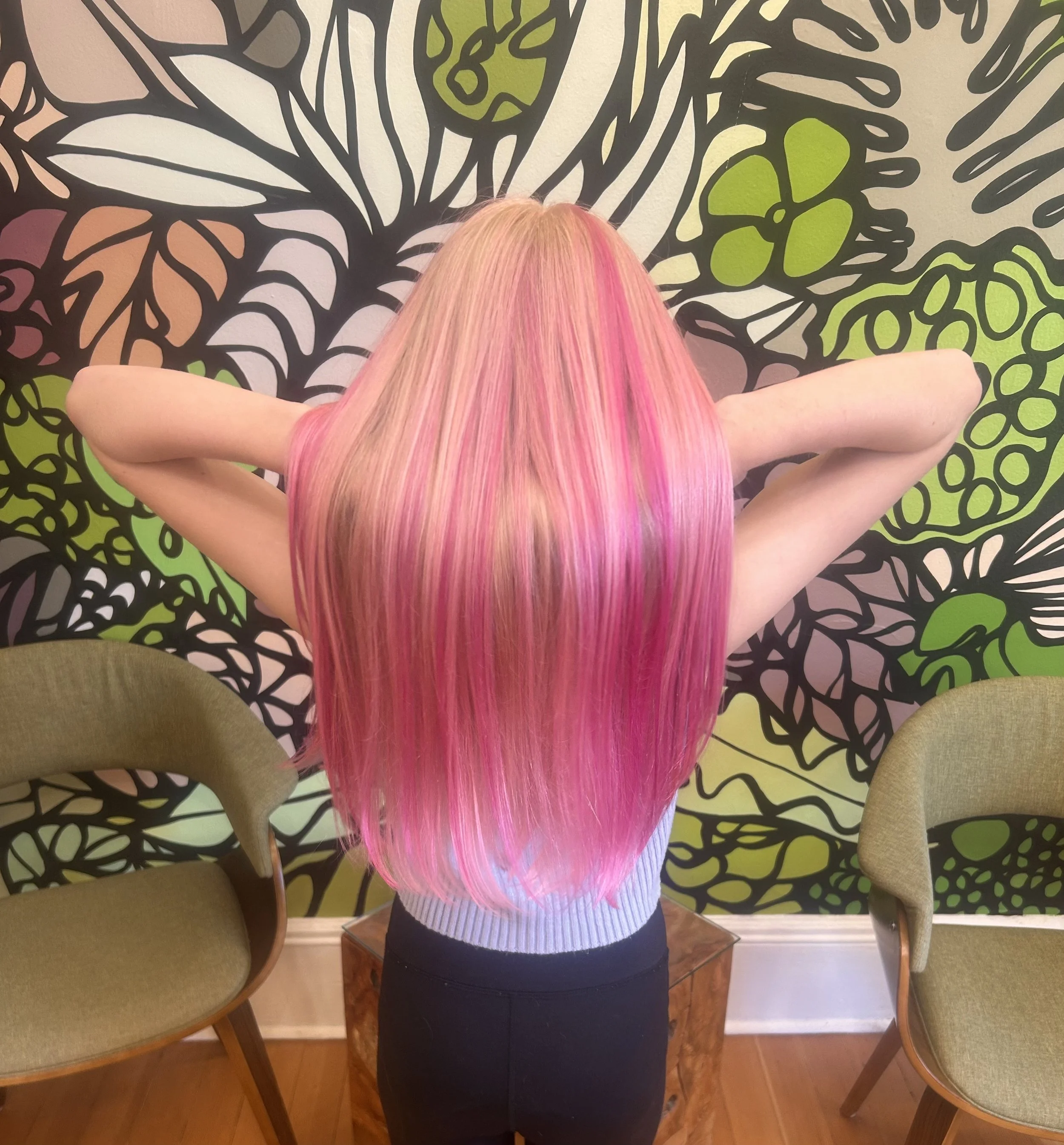 Back of a woman with long, straight hair that is dyed in shades of pink and blonde, sitting at a table in front of a mural with black outlines of various plant leaves and flowers, wearing a white ribbed sweater and dark pants.