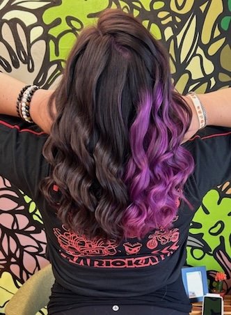 A person with long wavy brown and purple hair, back facing, with a colorful patterned wall in the background.