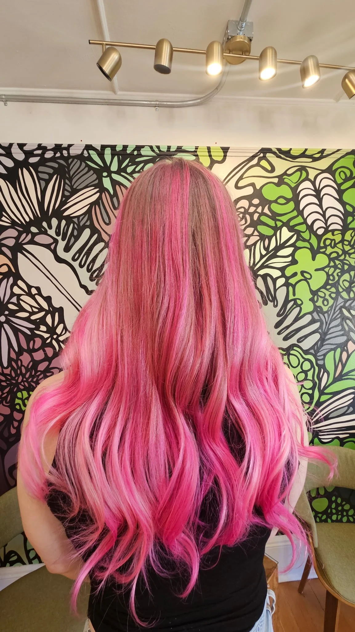 Person with long, wavy bright pink hair viewed from behind in front of colorful botanical wallpaper