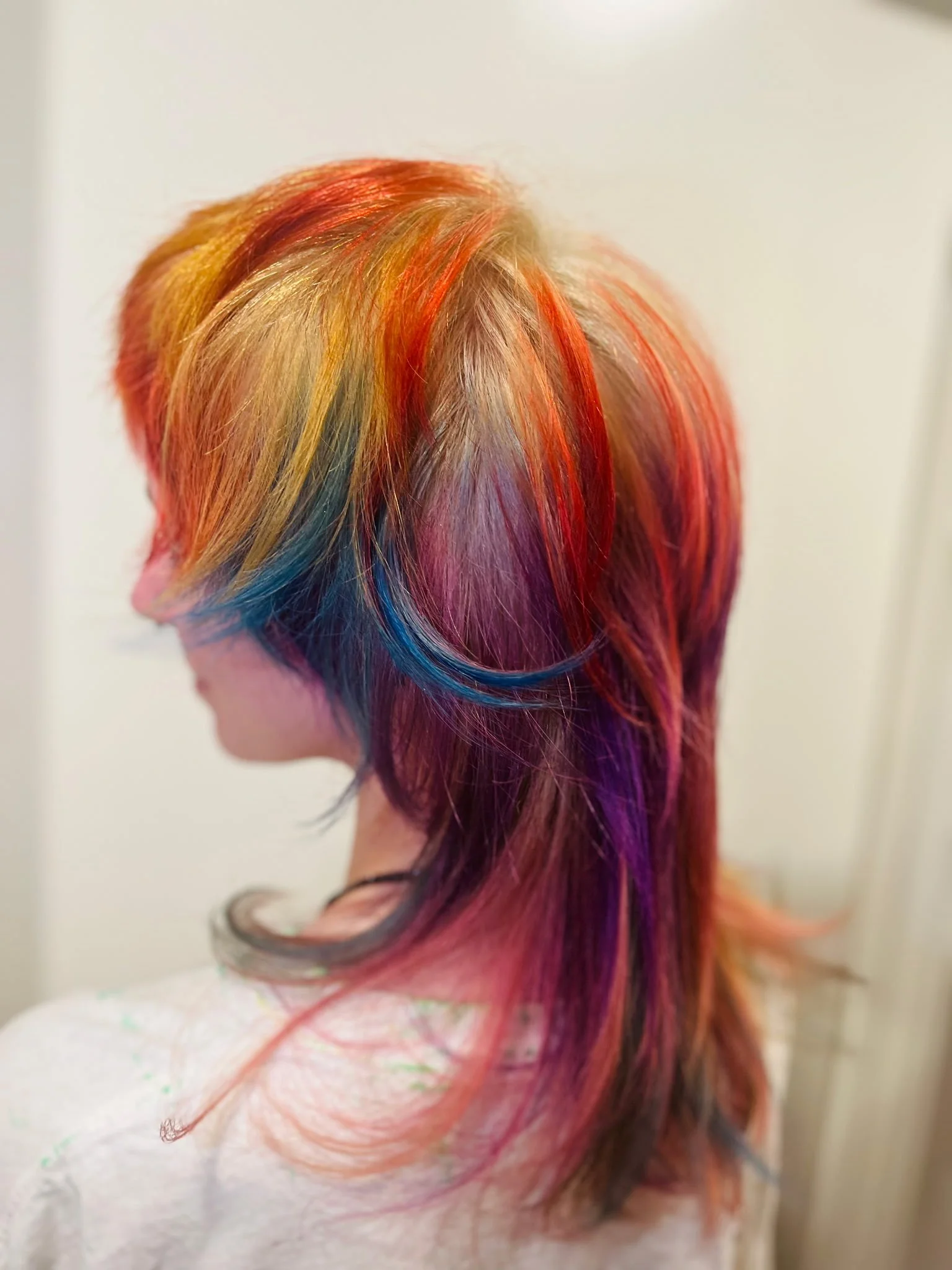 Close-up of a person's head with multi-colored, rainbow hair, showcasing shades of red, orange, yellow, green, blue, and purple, styled in loose waves.