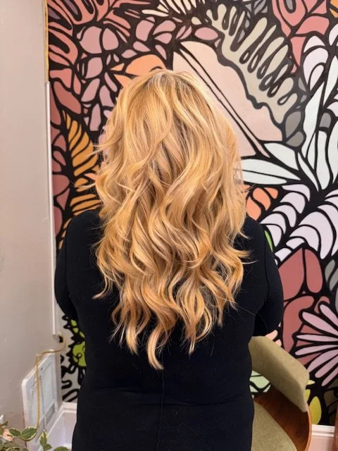 Back view of a woman with long, wavy blonde hair, wearing a black top, standing in front of a colorful, abstract mural.