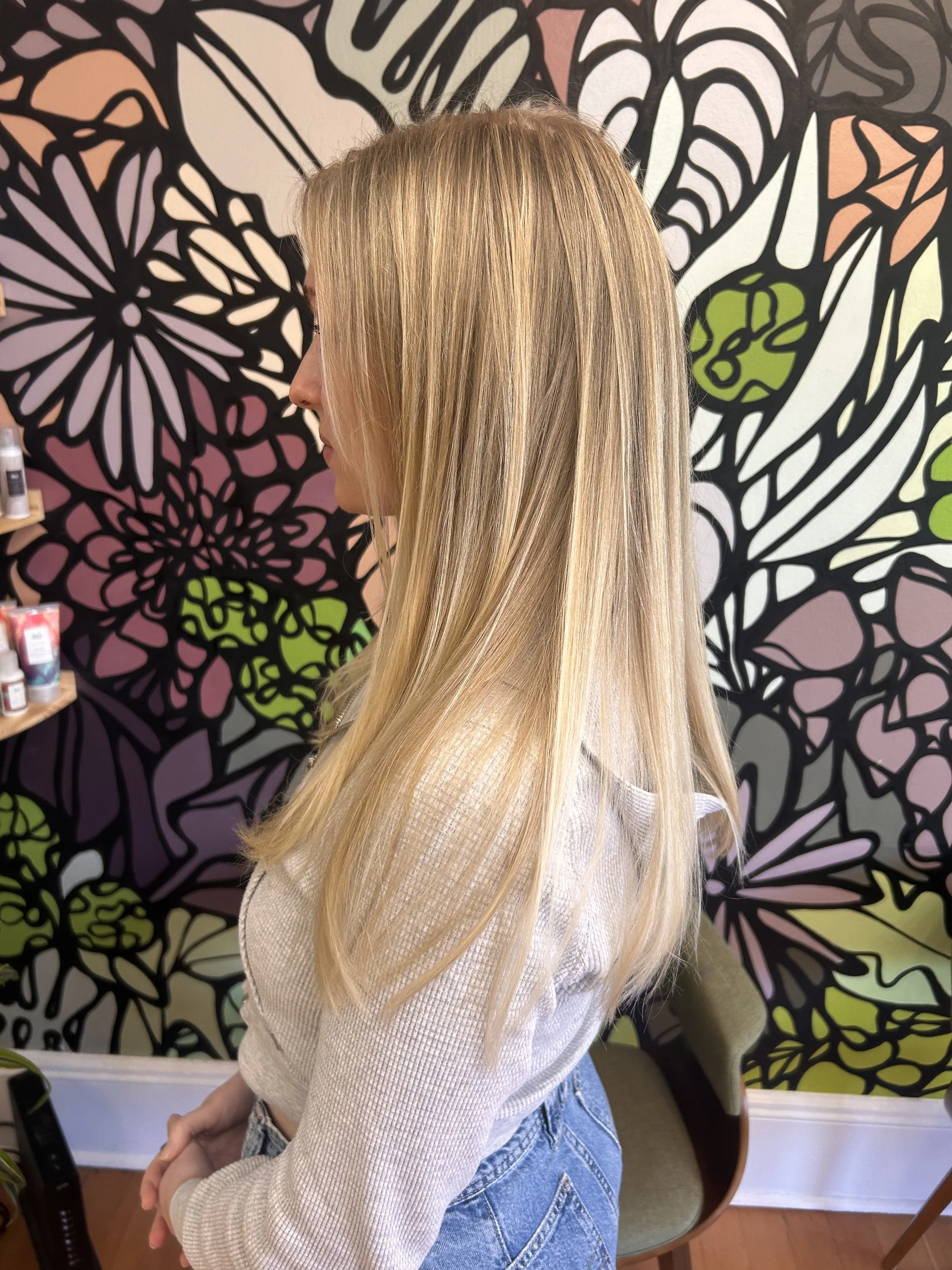 Woman with long blonde hair wearing a white sweater and jeans, seated in front of colorful floral wallpaper