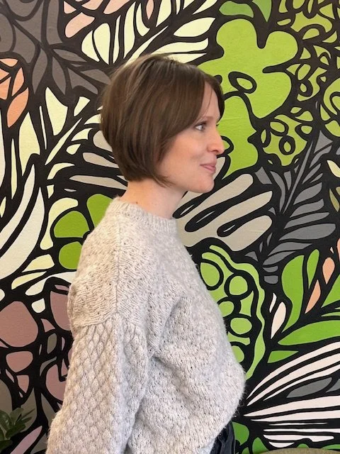 Side-profile portrait of a woman in a white knit sweater standing before a colorful abstract mural.