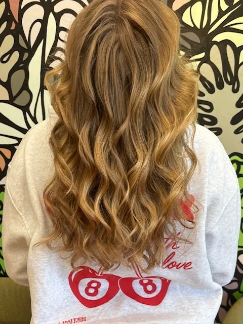 A woman with long, wavy blonde hair, seen from behind, wearing a gray sweatshirt with red cherries and the word 'love' on it, standing against a decorative black and green floral wall.