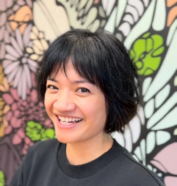 A person with short black hair smiling in front of a colorful floral mural.