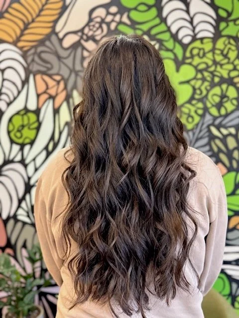 Back of a woman with long, wavy brown hair standing in front of a colorful mural with abstract floral designs.