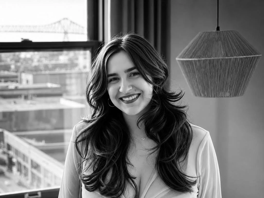 Smiling woman with long wavy hair posing indoors beside a hanging pendant lamp and city-view window.