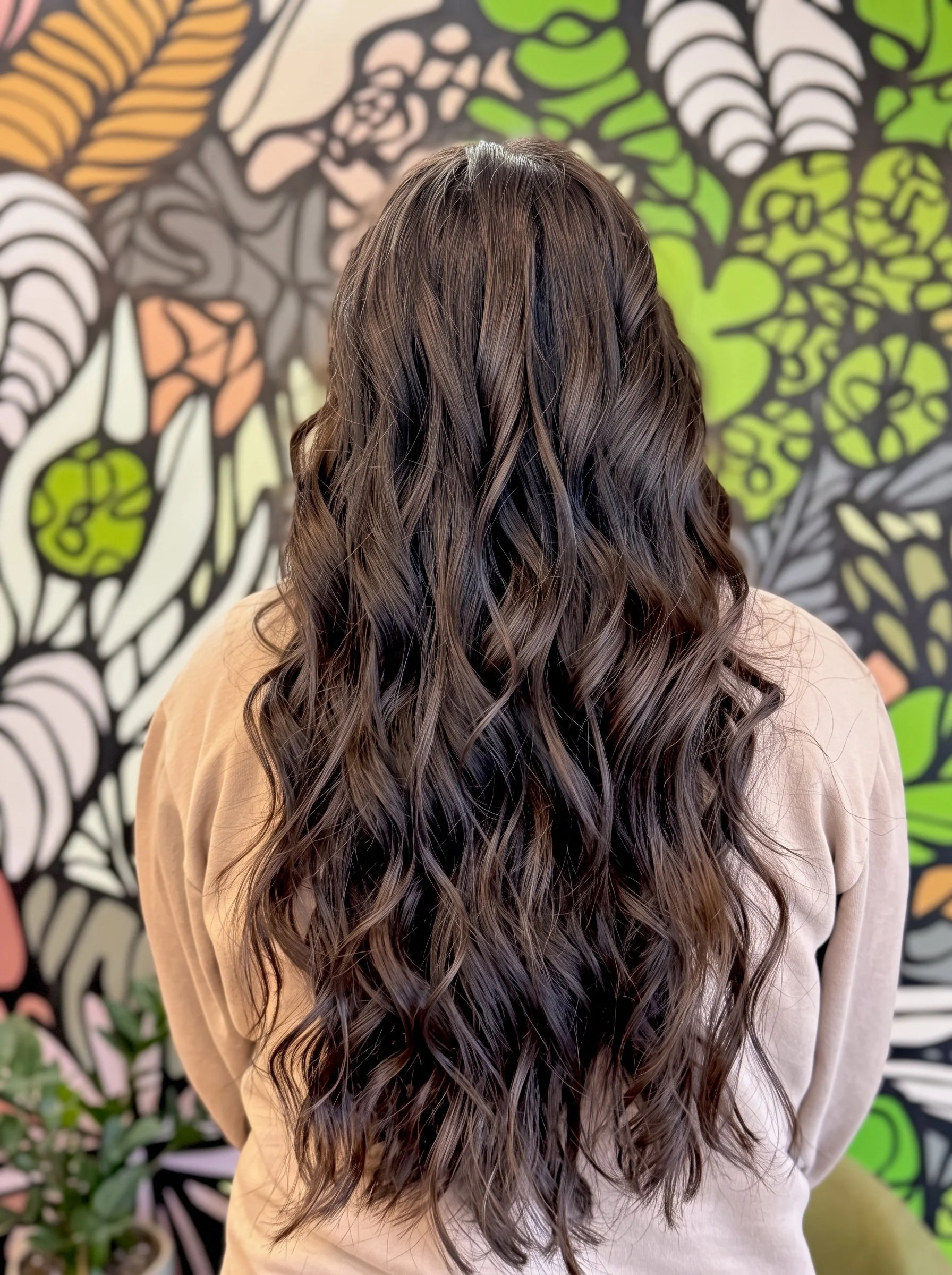 Back view of a person with long, dark brown wavy hair against a colorful botanical mural