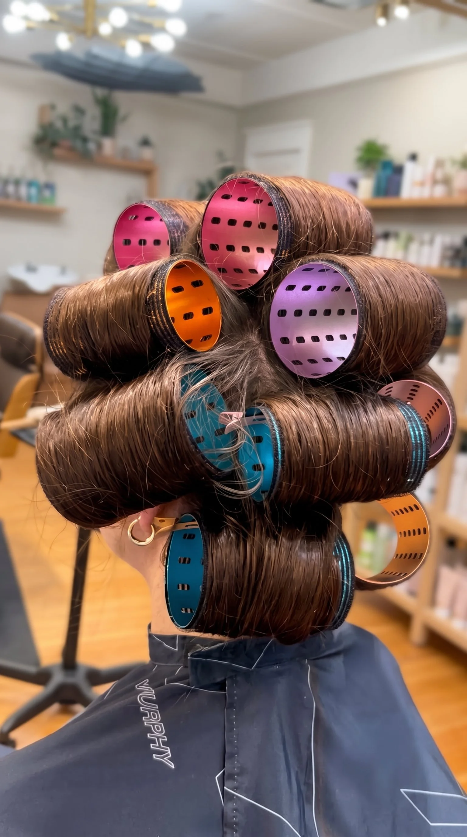 Back view of a client wearing multiple colorful hair rollers in a salon chair.
