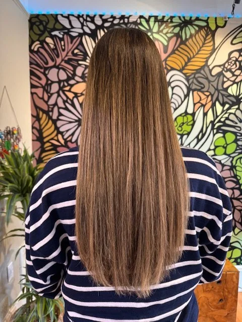 Back view of a woman with long, straight brown hair, wearing a navy blue and white striped sweater, standing indoors with a colorful floral and leaf pattern mural behind her.