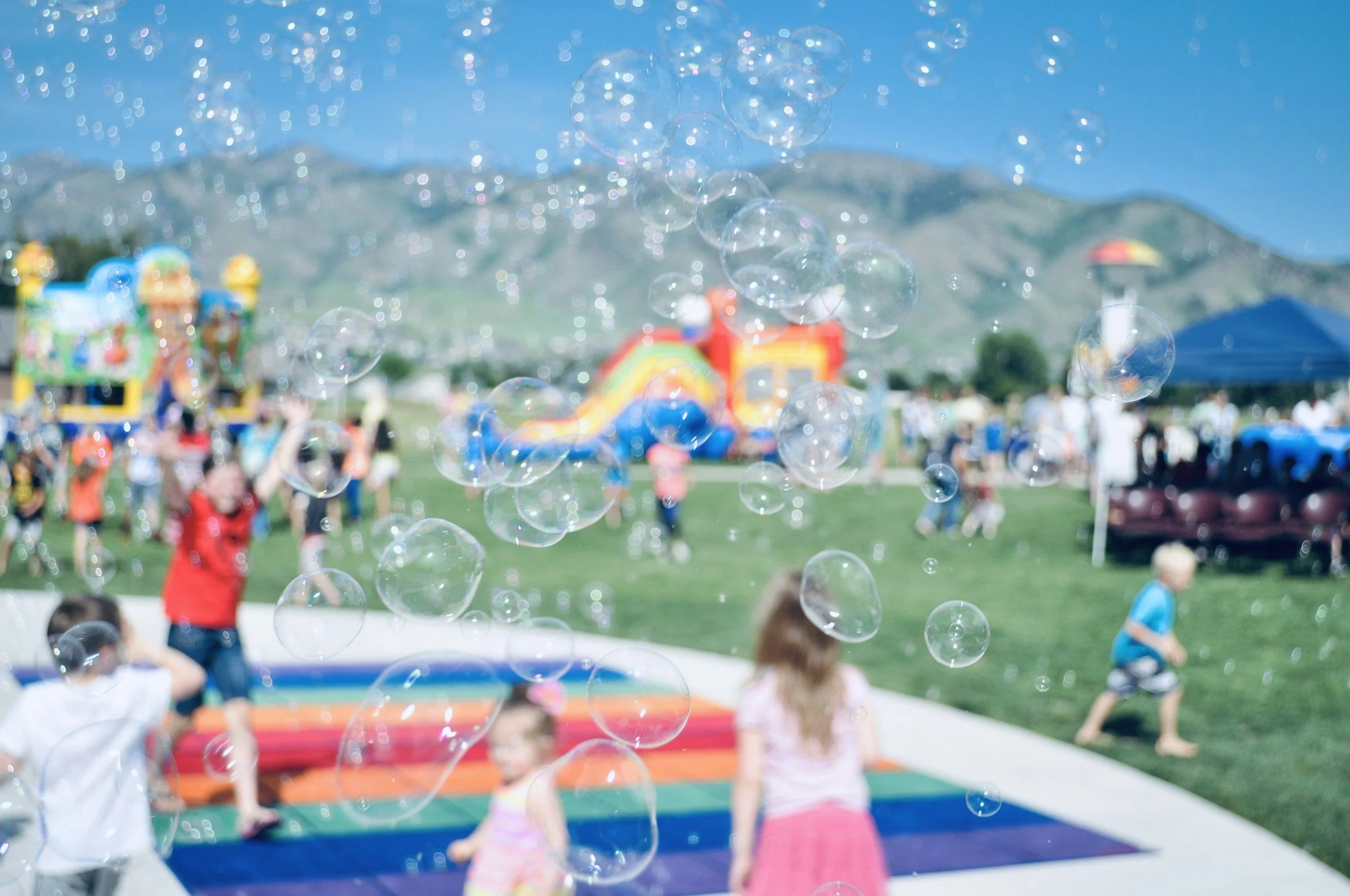 Children playing with bubbles at a colorful outdoor festival with rainbow-colored ground, inflatable bounce house, and a mountain backdrop