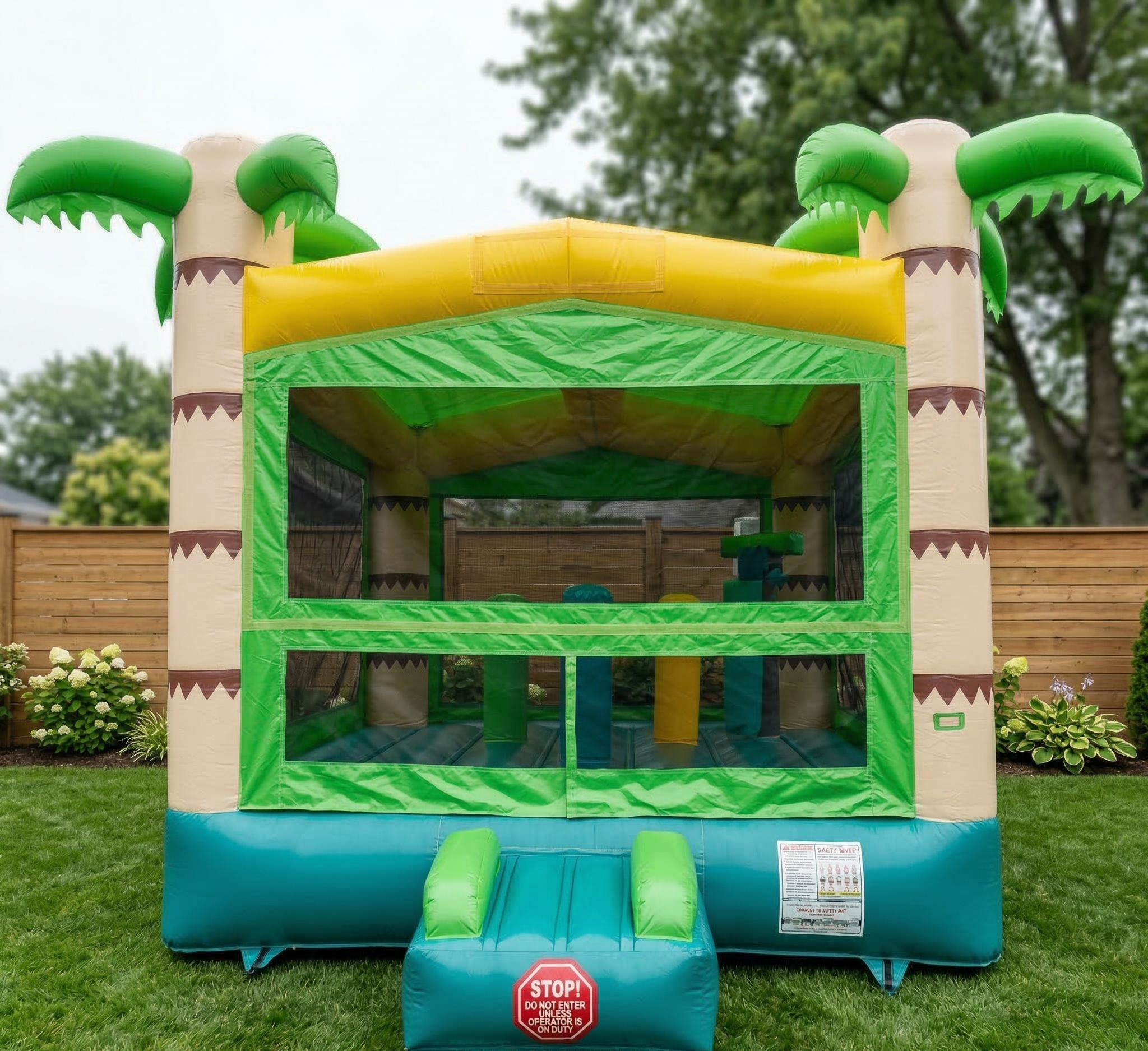 Colorful inflatable bounce house with palm tree designs, set in a backyard with a wooden fence and green grass, and some plants and flowers in the background.