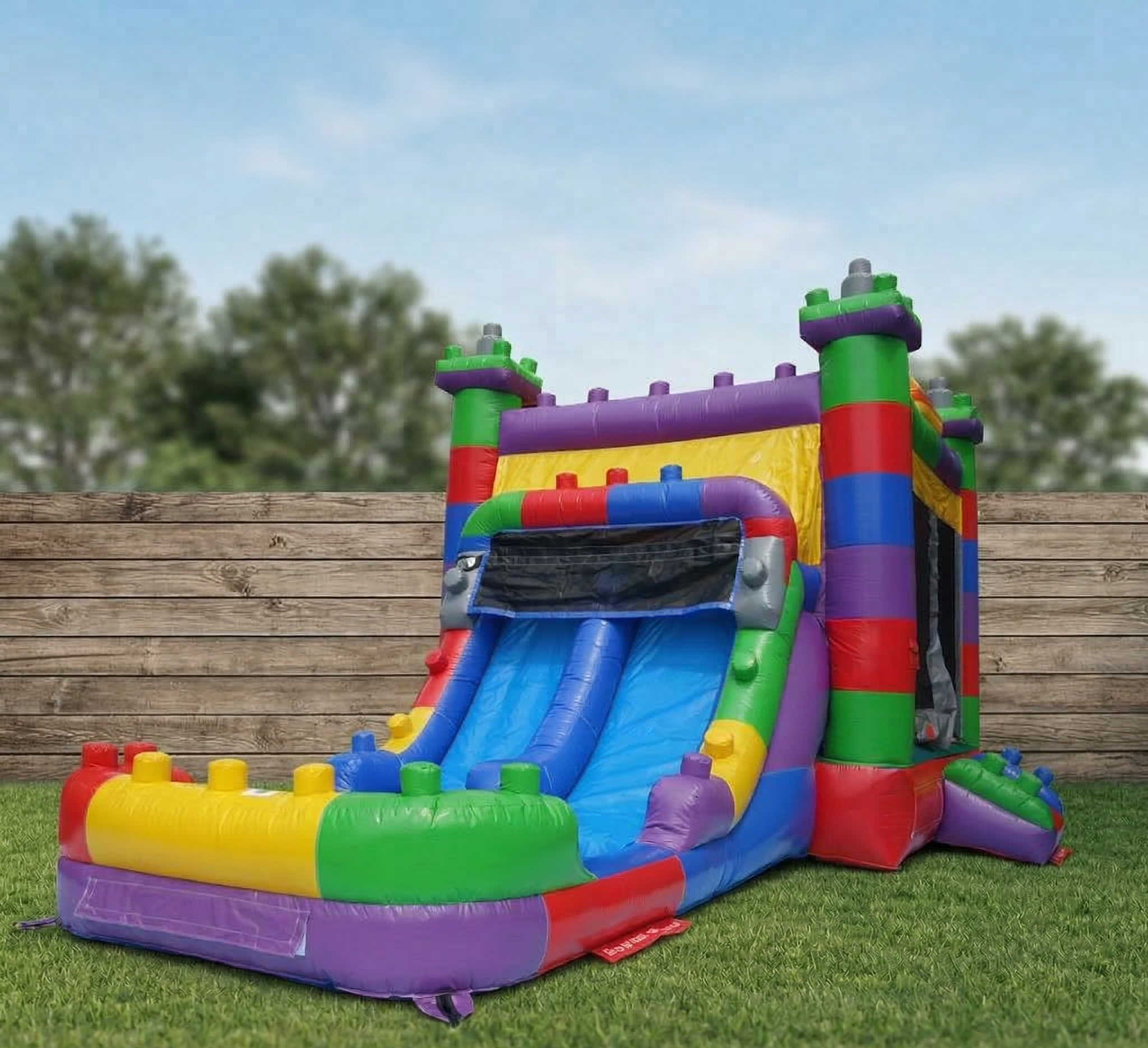 Colorful inflatable bounce house with slides, set up outdoors on grass with a wooden fence and trees in the background.