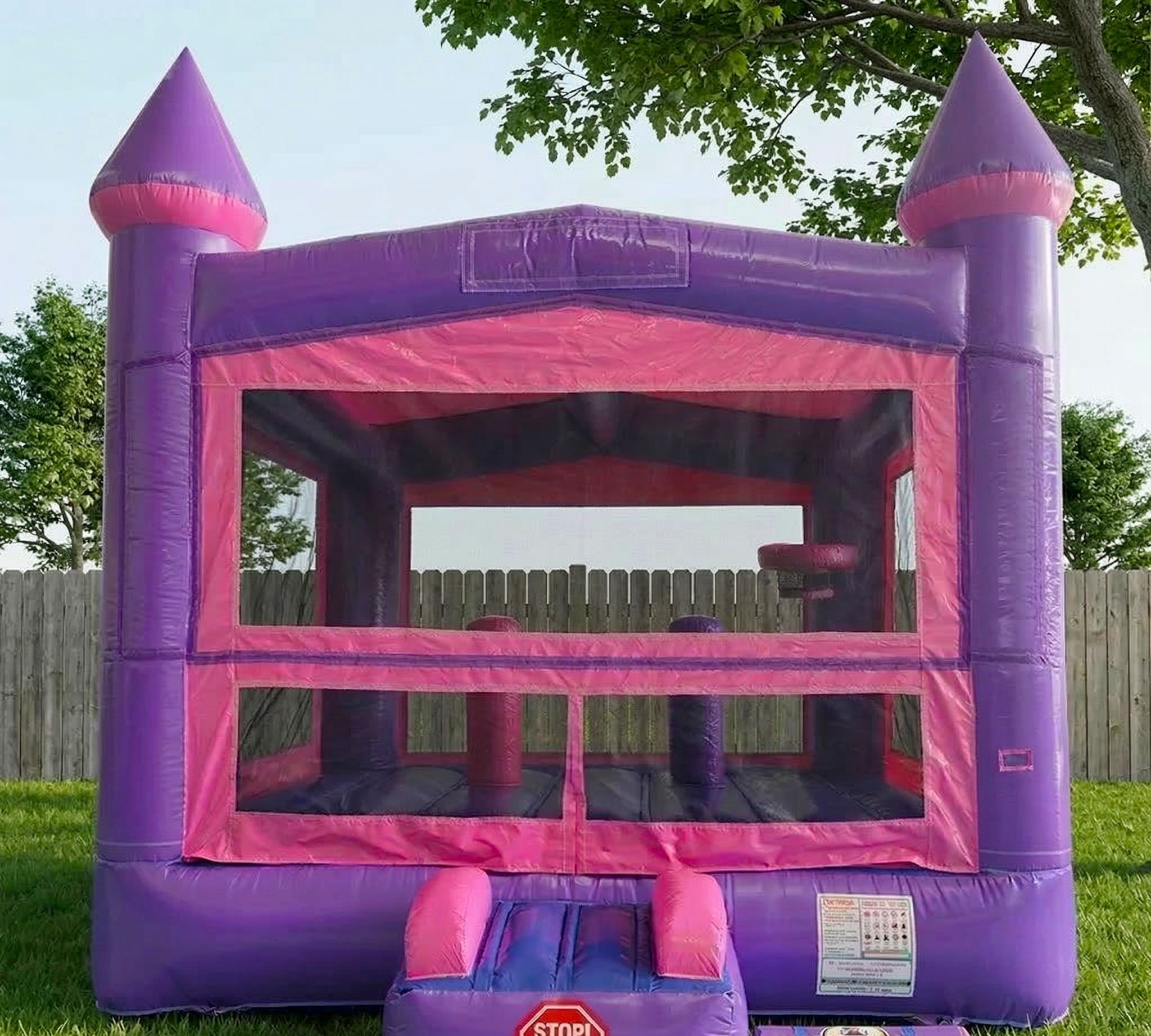 Pink and purple inflatable castle with a small entrance slide, set up on a grassy yard behind a wooden fence, with trees and a clear sky in the background.