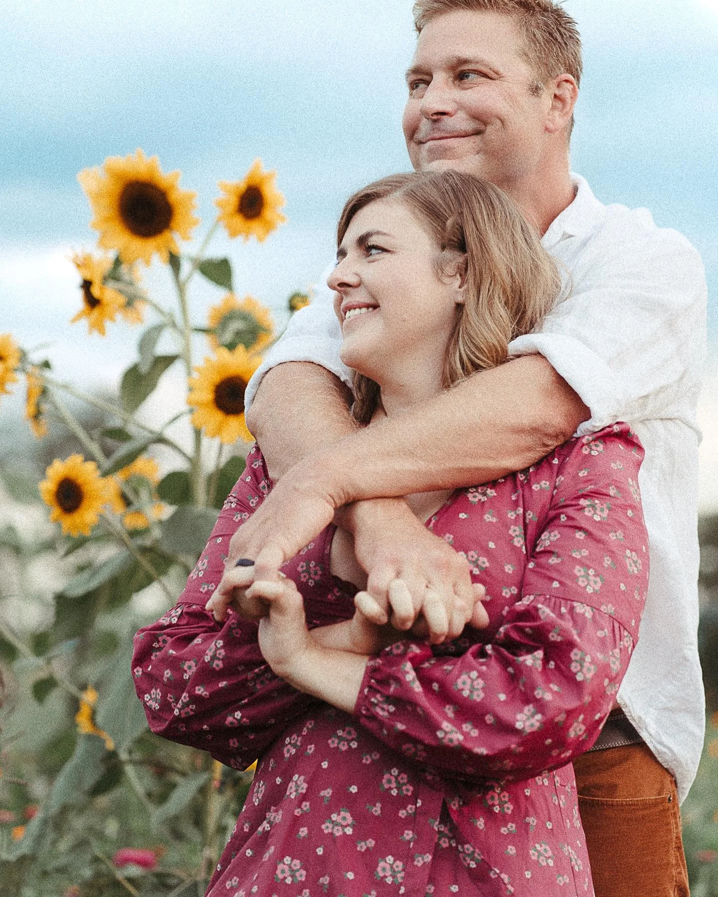 A couple stands in a sunflower field, with the man embracing the woman from behind, both smiling and looking happy.
