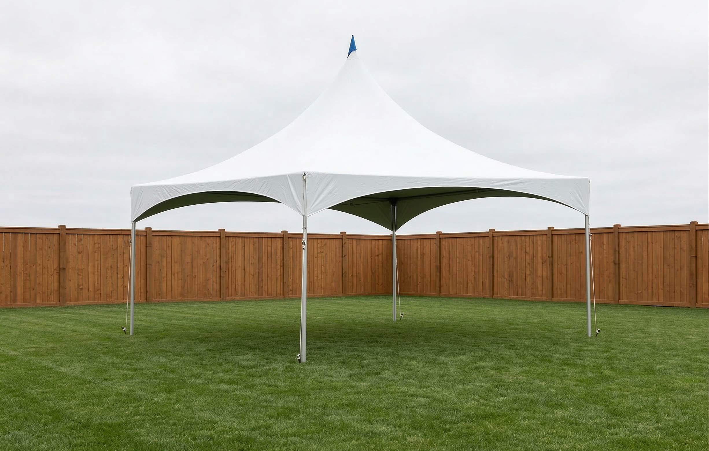A large white outdoor canopy tent set up on a grassy backyard with a brown wooden fence in the background and cloudy sky overhead.