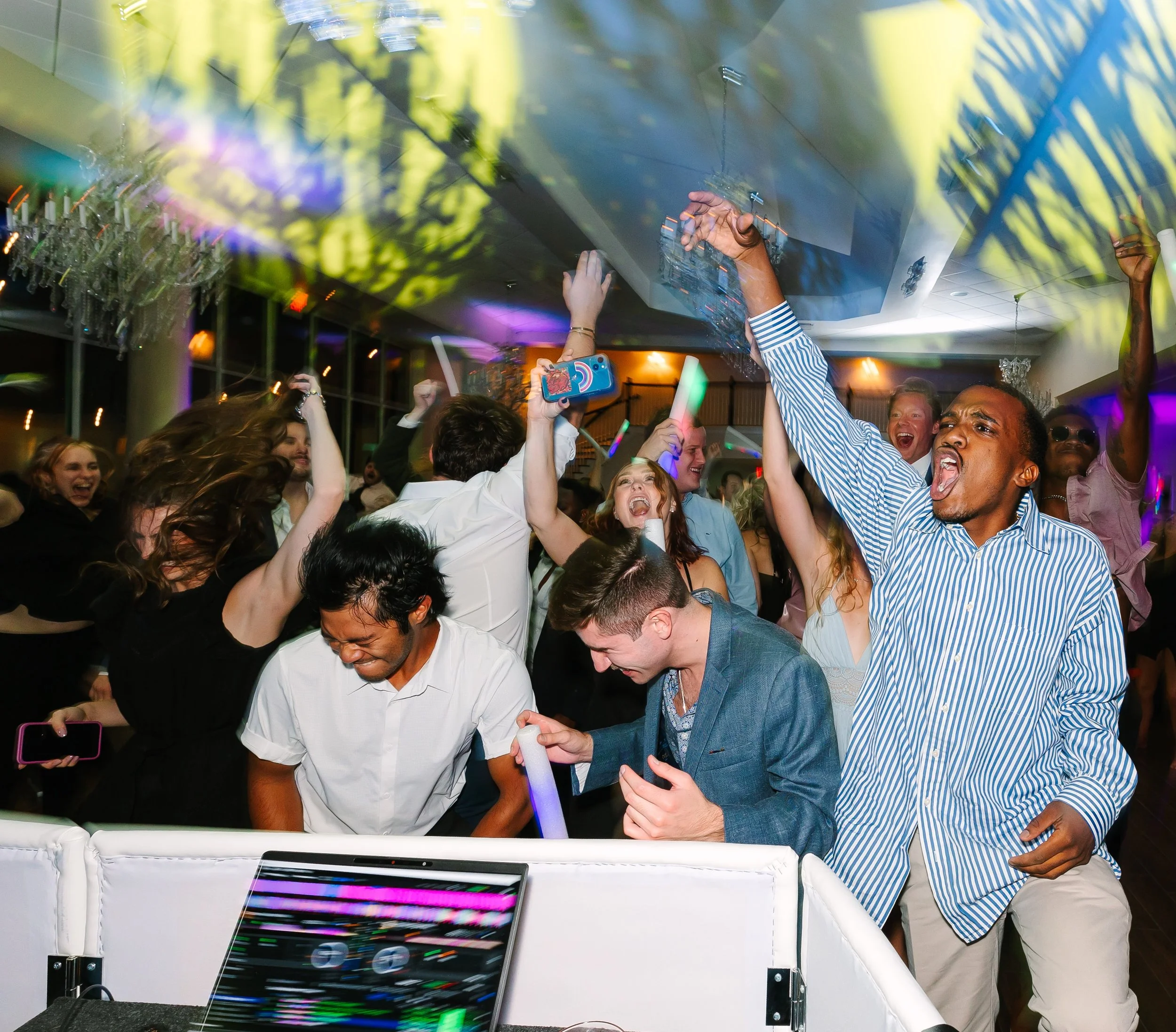 People dancing and having fun at a party with colorful lights, some holding glow sticks, in an indoor setting with chandeliers and modern decor.