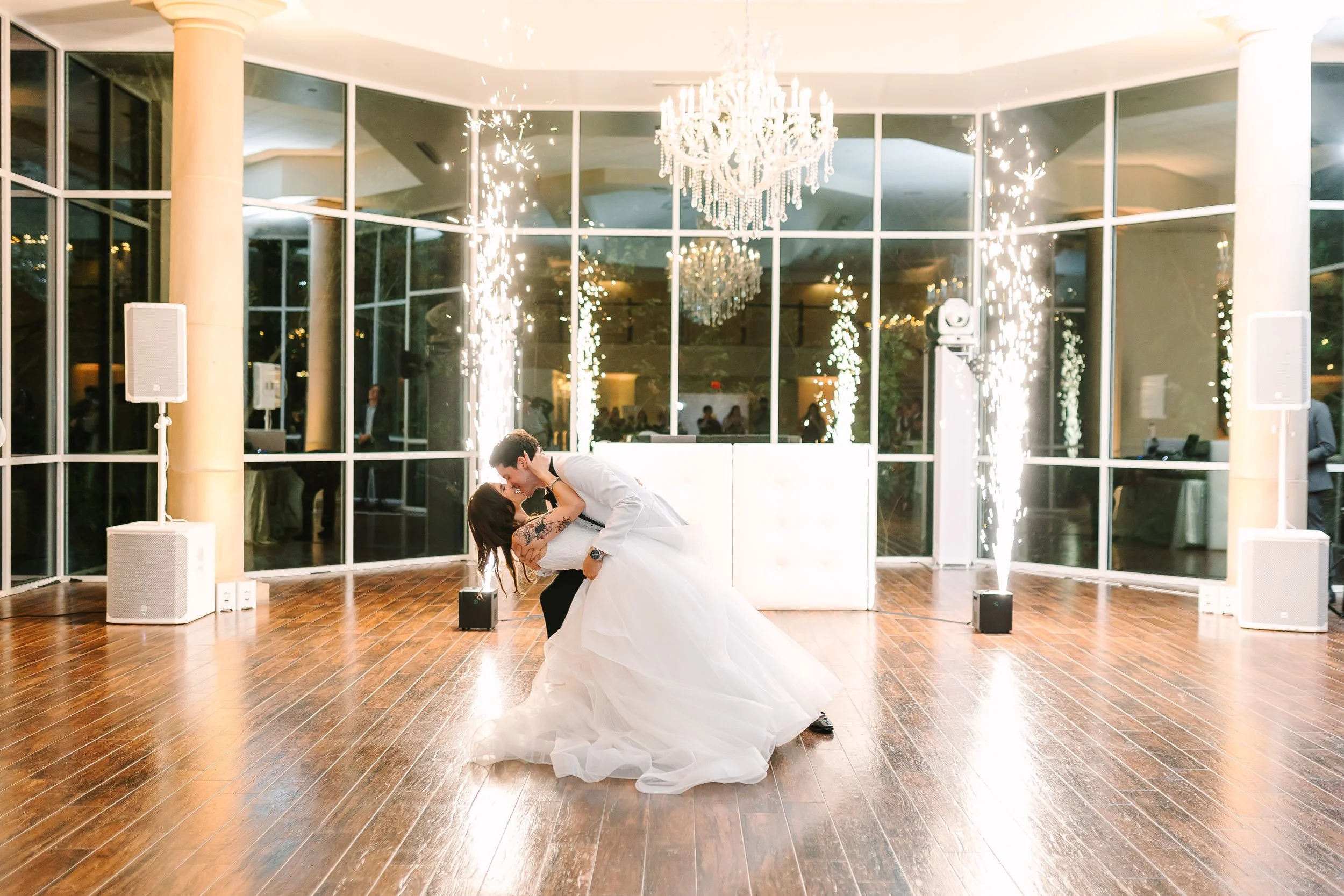 A newly married couple is sharing a kiss and dance at their wedding reception inside a modern venue with large glass windows, chandeliers, and sparklers.