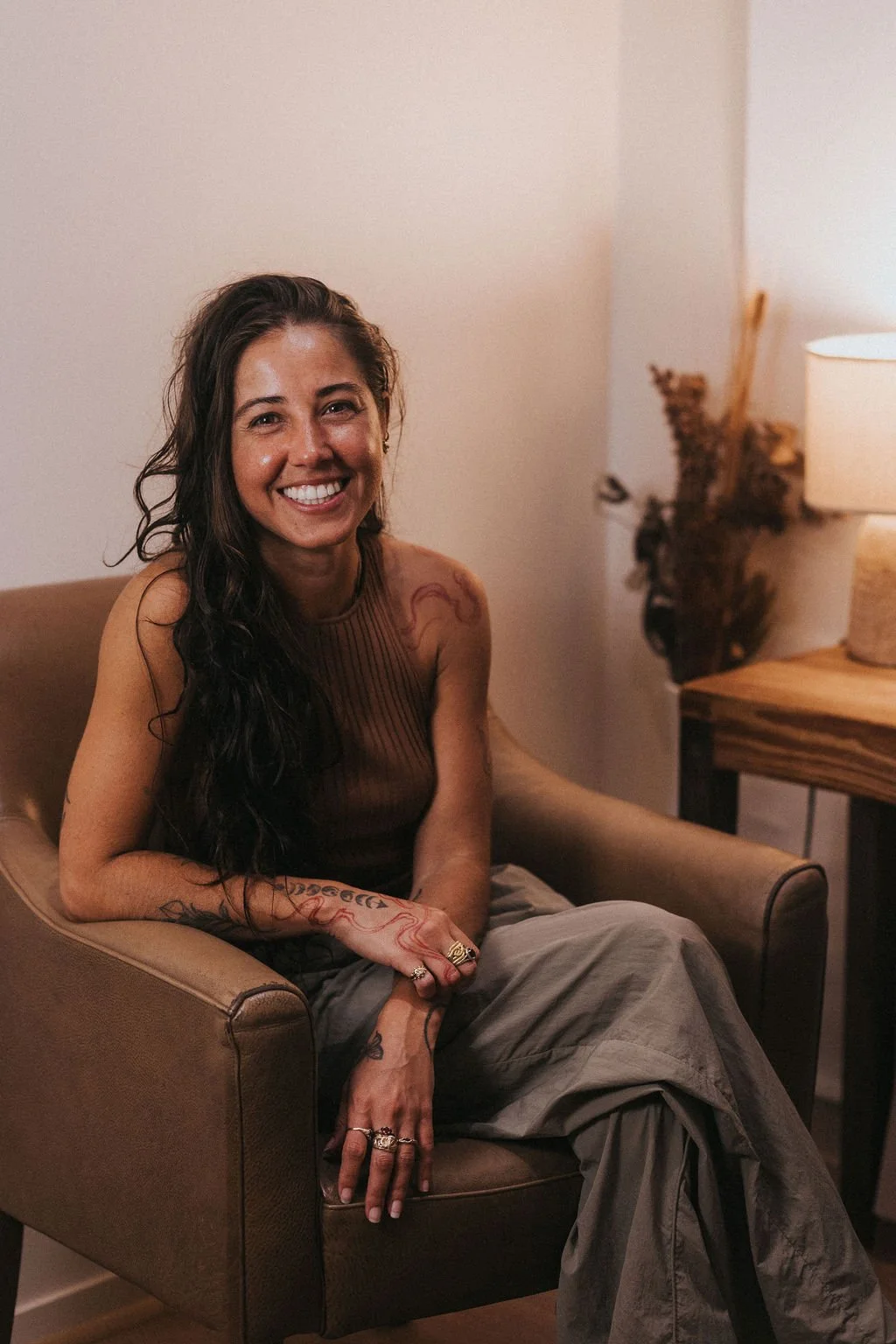 A smiling woman with long dark hair, wearing a sleeveless brown top and beige pants, sitting on a brown armchair in a warmly lit room with a wooden side table and a lamp.