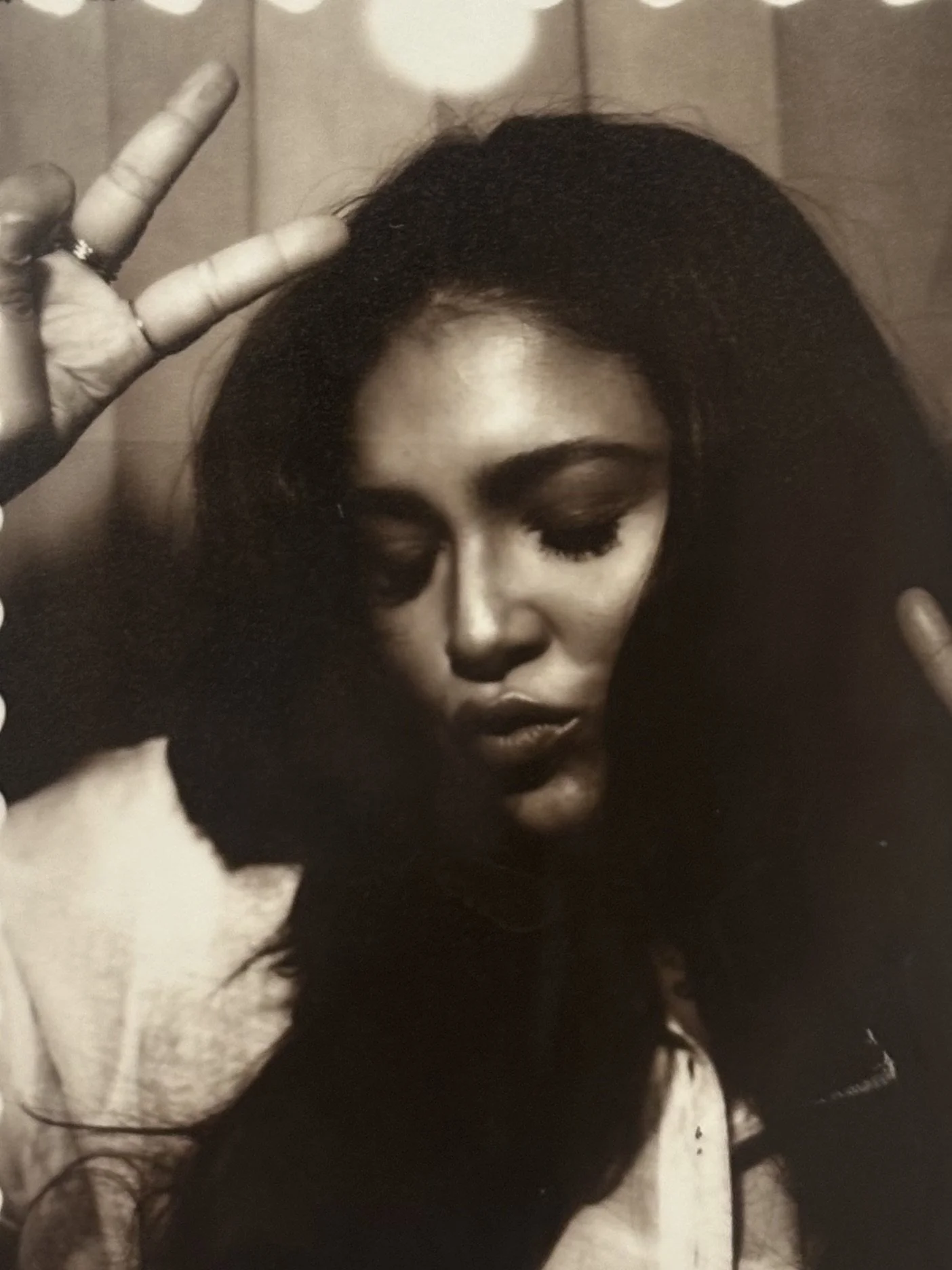 A woman with dark hair and closed eyes making a peace sign with her hand. Black and white photo with a blurry background.