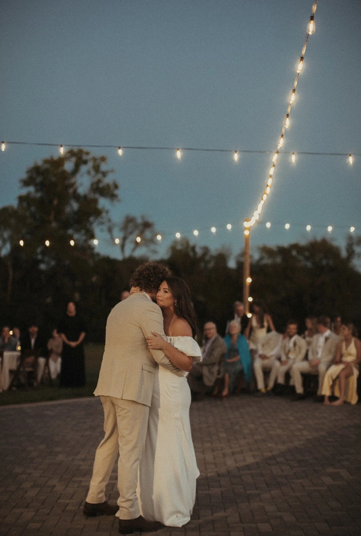 At an outdoor evening wedding reception under string lights, a bride and groom dance closely together with guests seated in the background.