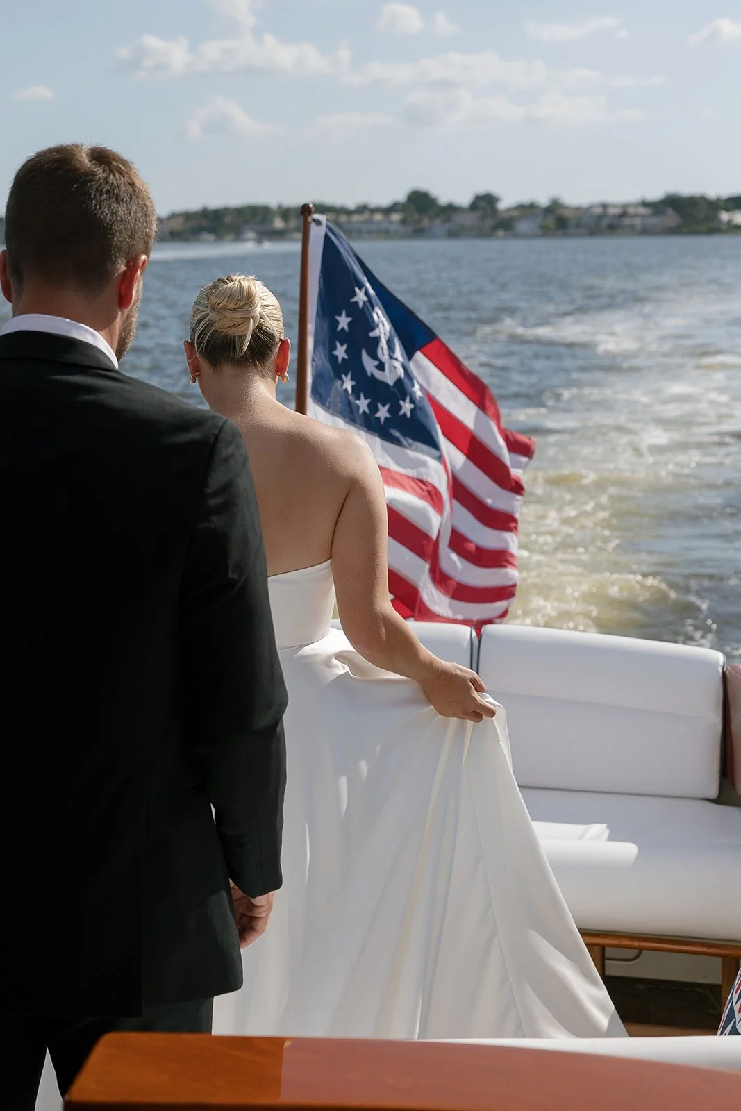 A couple dressed in formal wedding attire on a boat with an American flag, as they hold hands and look toward the water on a sunny day.