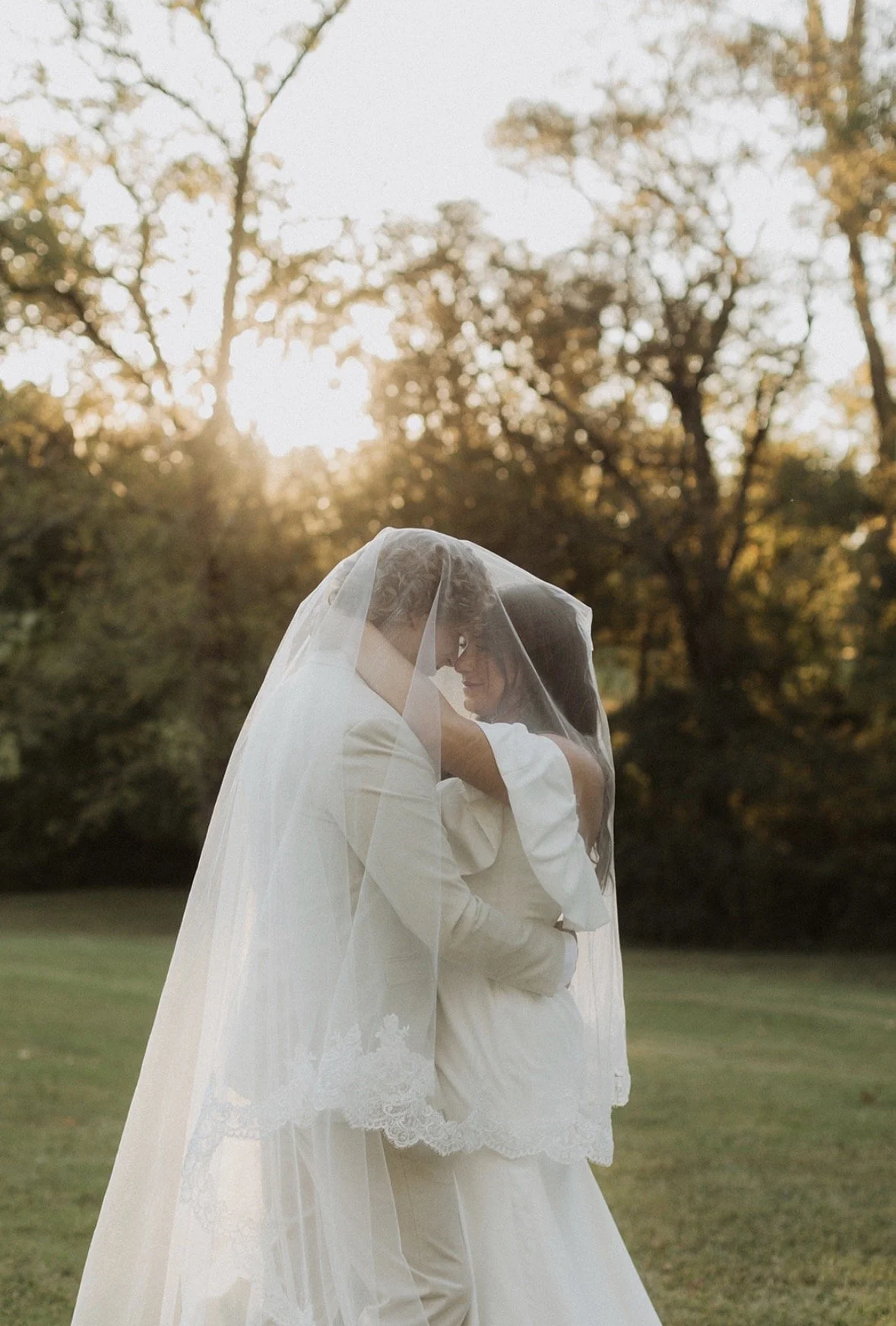 A bride and groom embrace outdoors, the bride wearing a veil and wedding dress, the groom in a suit, with autumn trees and sunlight in the background.