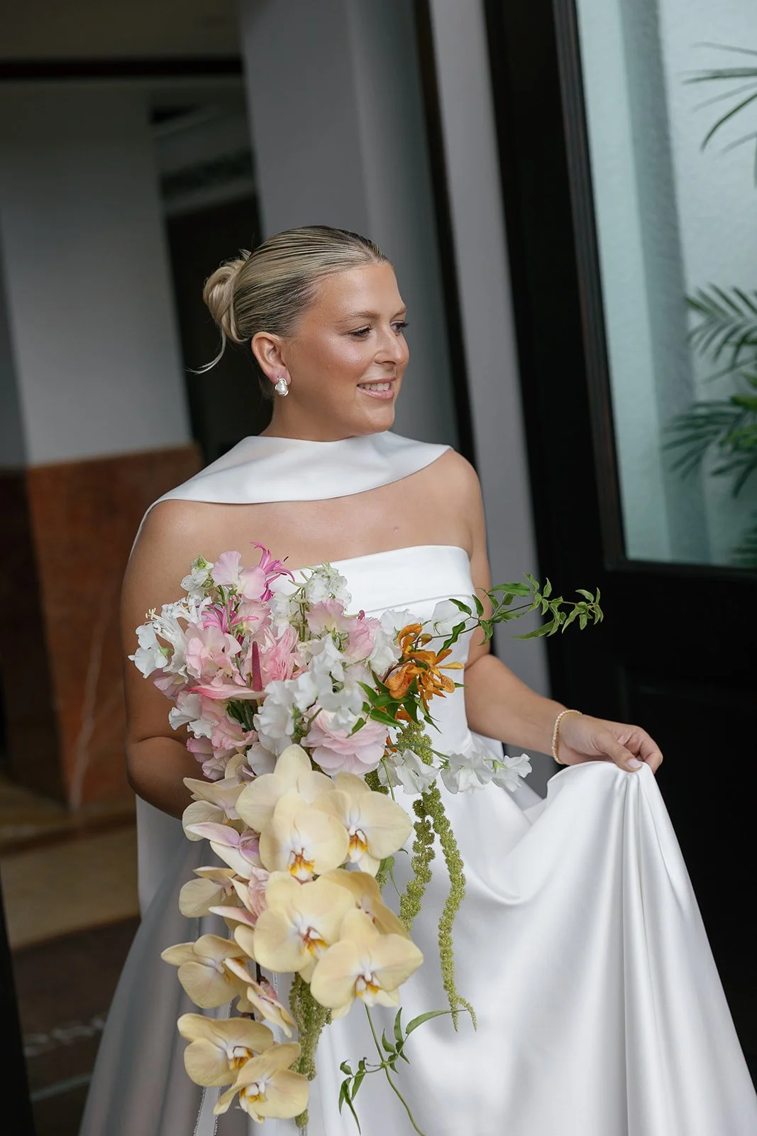 A bride in a white wedding dress holding a bouquet of pink, white, and yellow flowers, looking out a window.