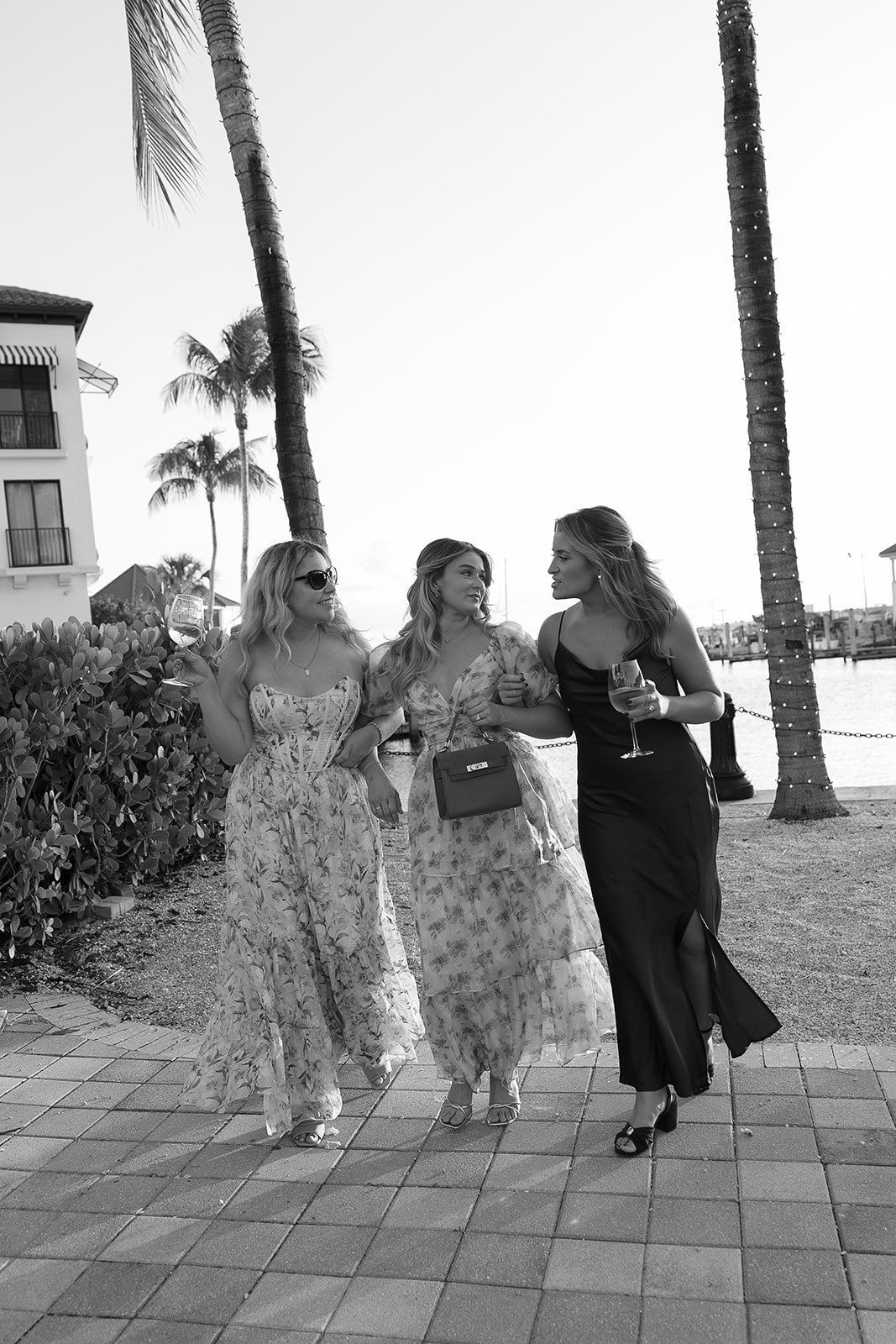 Three women in elegant dresses walking and talking along a waterfront with palm trees, buildings, and string lights in the background, holding glasses of wine.