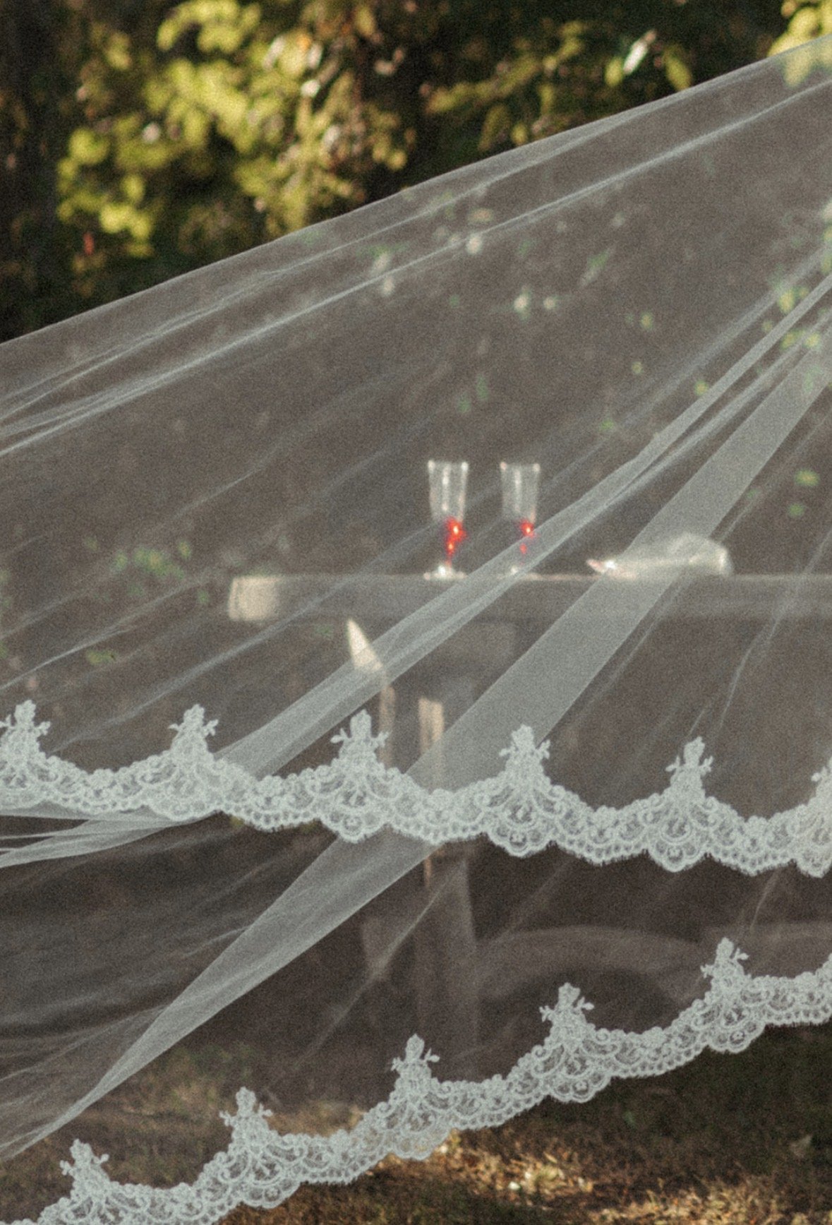 Two glasses with red wine sitting on a table covered by a lace tablecloth, with a sheer fabric draped over the table, and a background of green foliage.