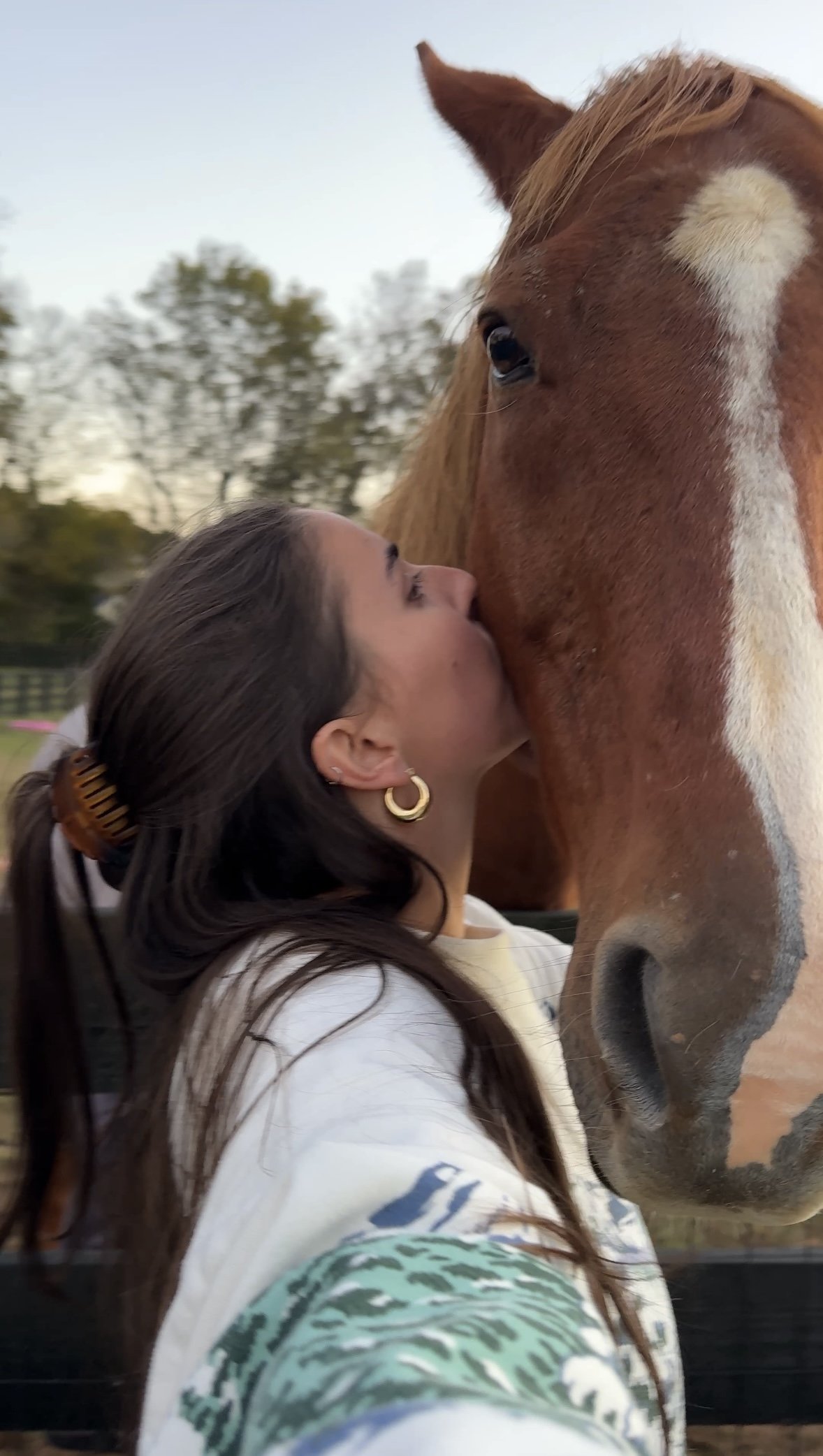 A woman with long dark hair, gold hoop earrings, and a white jacket with green and blue patterns, is kissing a large brown and white horse on its face outdoors during daytime.