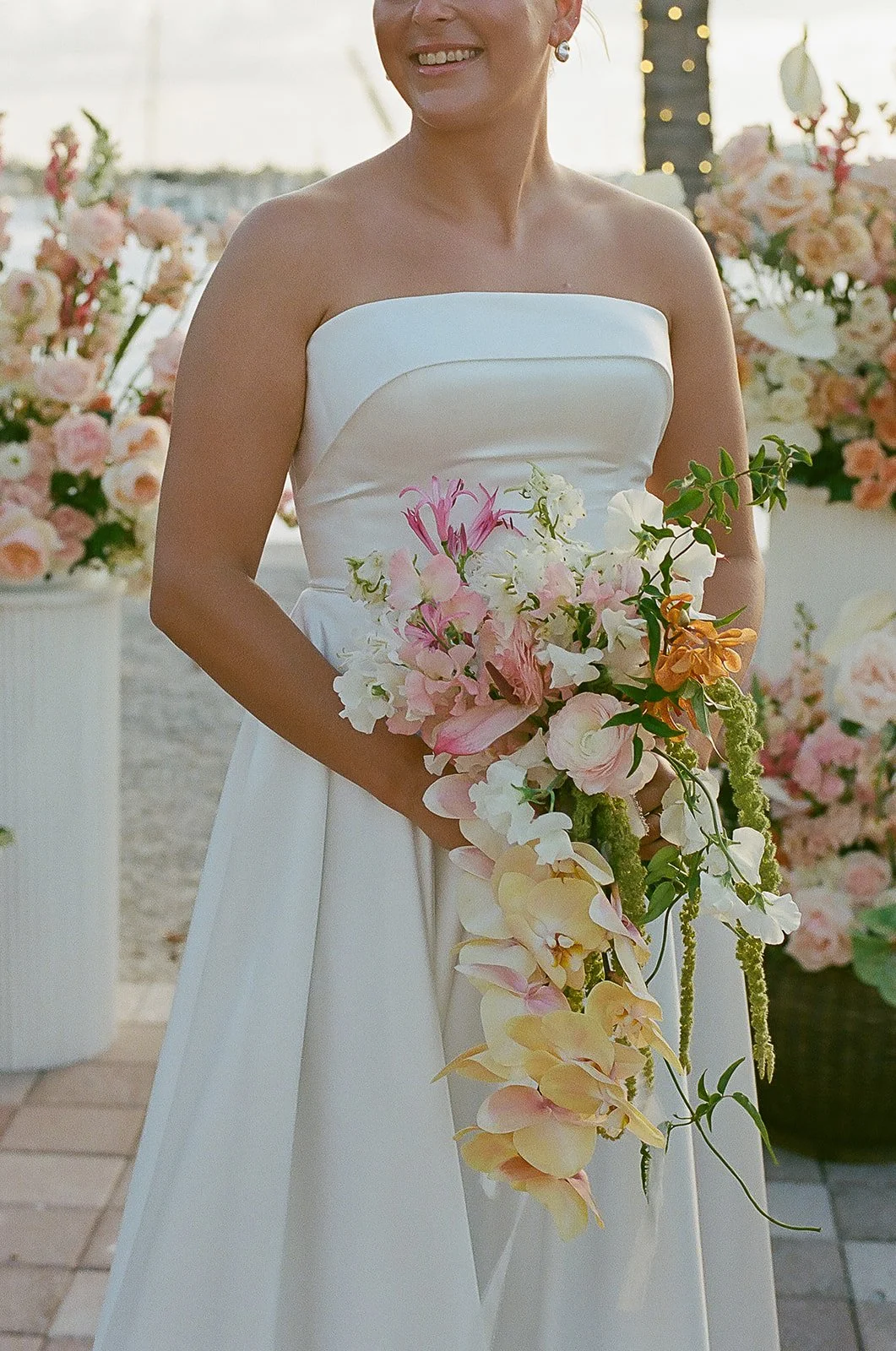 Woman in white strapless wedding dress holding a cascading bouquet of pink, white, peach, and yellow flowers at an outdoor wedding.