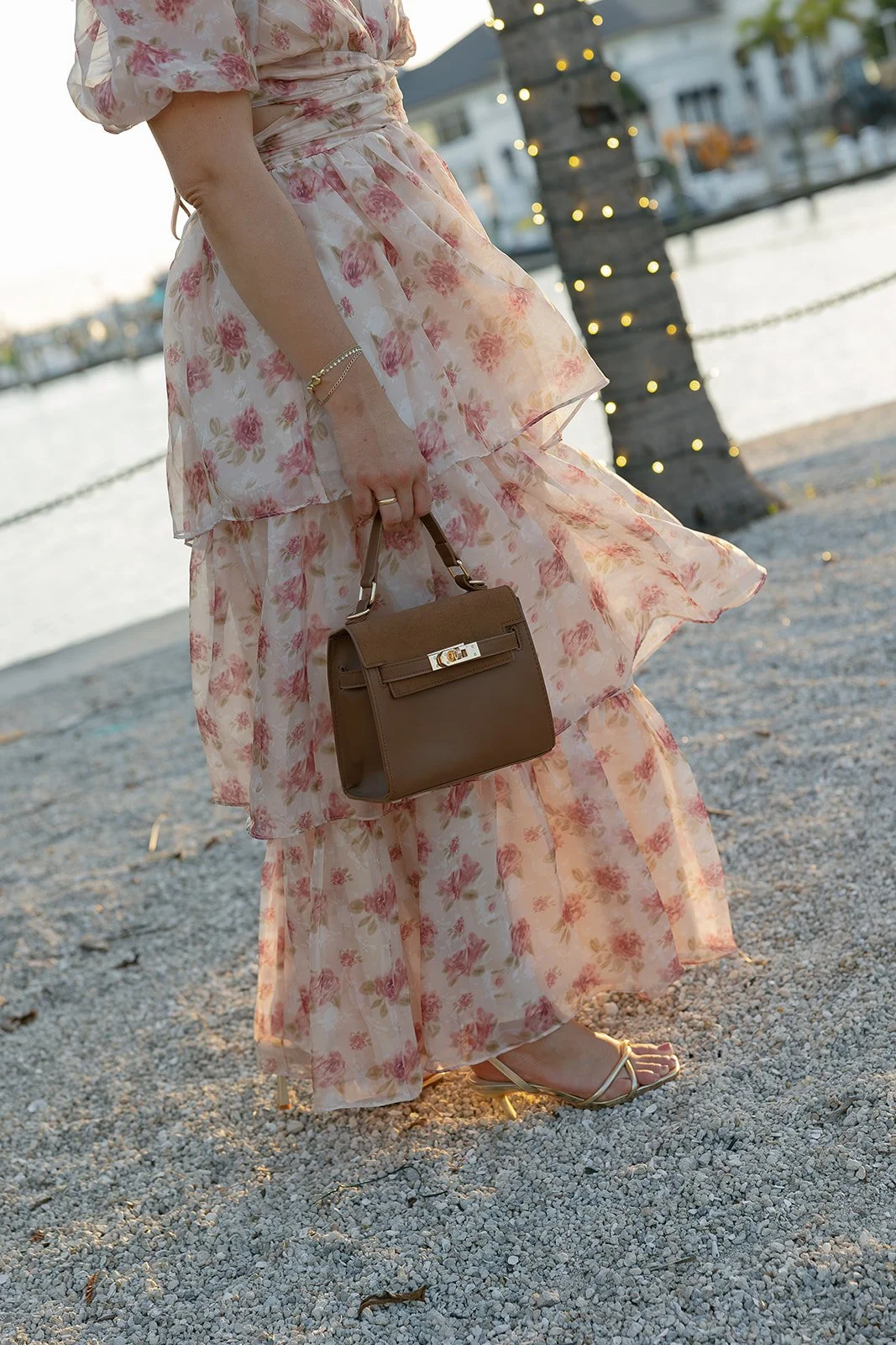 A woman in a floral maxi dress and gold sandals holding a brown handbag standing on a gravel beach with a lighthouse and boats in the background.