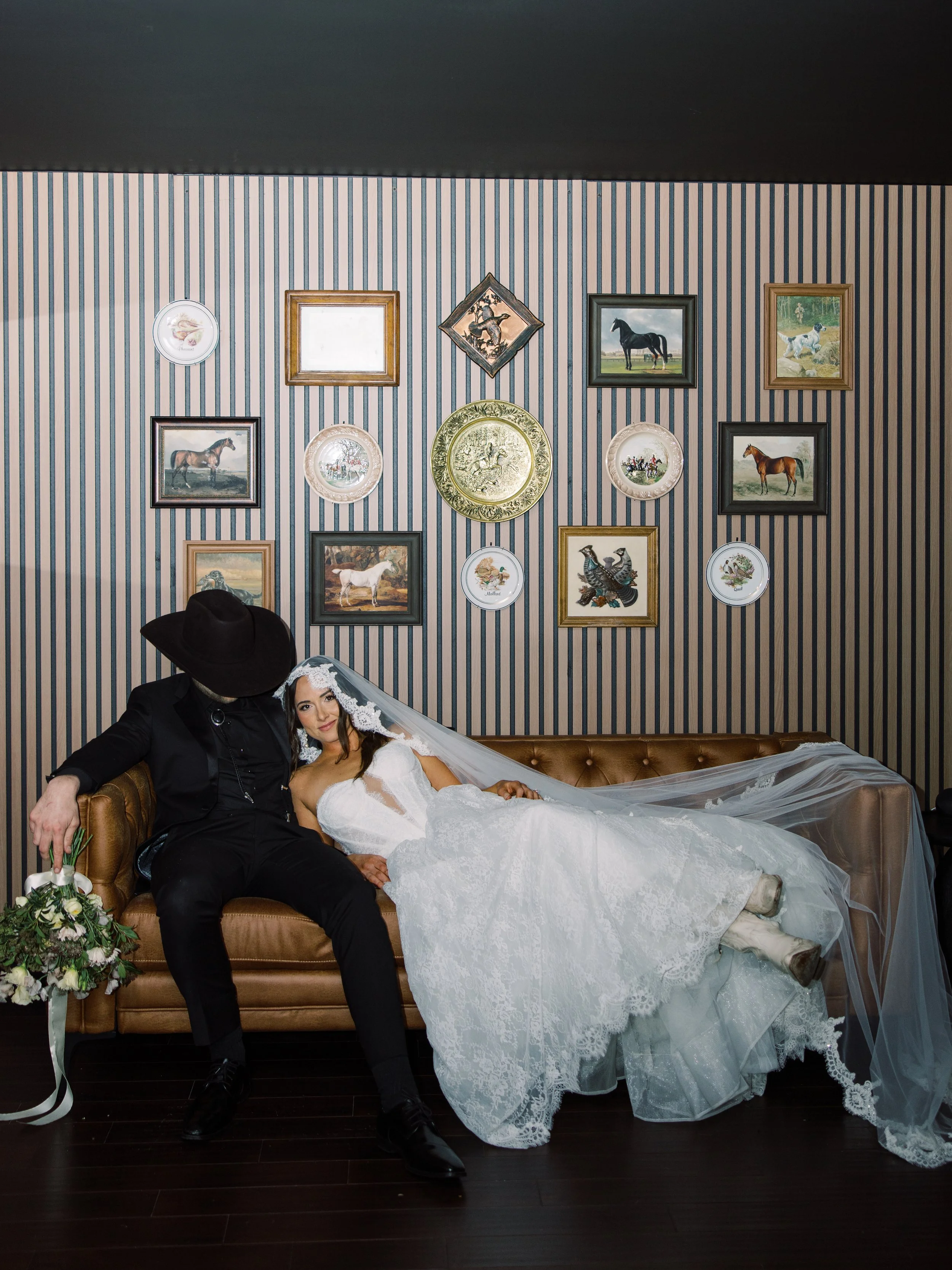 A bride in a white wedding dress and veil lying on a brown tufted sofa with a man in dark clothing and a wide-brimmed hat, holding her hand, in a room with striped wallpaper and framed pictures of horses and other animals.