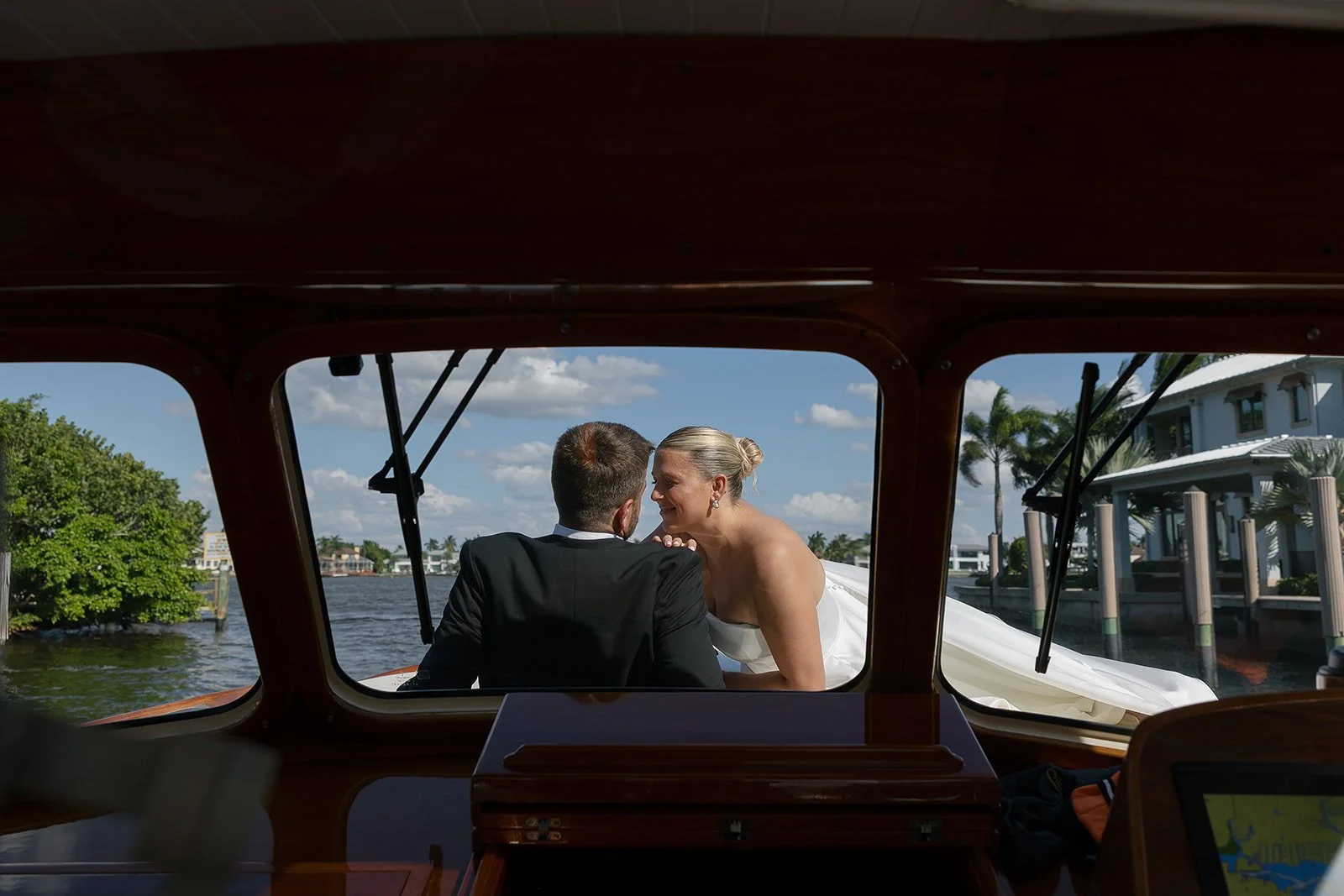 A couple celebrating their wedding and sharing a kiss on the deck of a boat, with a cityscape and water in the background.