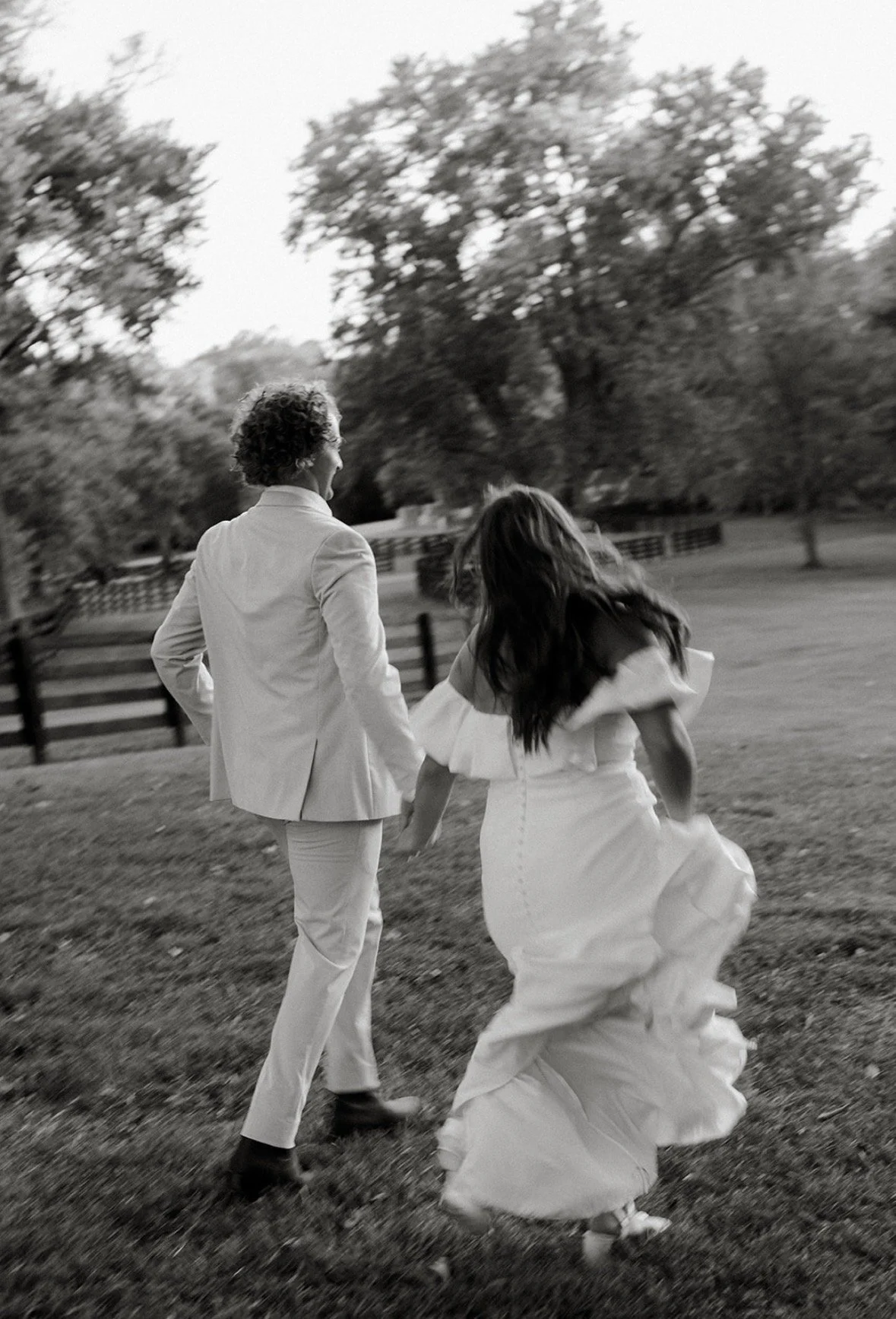 A black-and-white photo of a couple holding hands and running outdoors on grass, with trees and a fence in the background. The woman is wearing a long white dress, and the man is dressed in a light-colored suit.