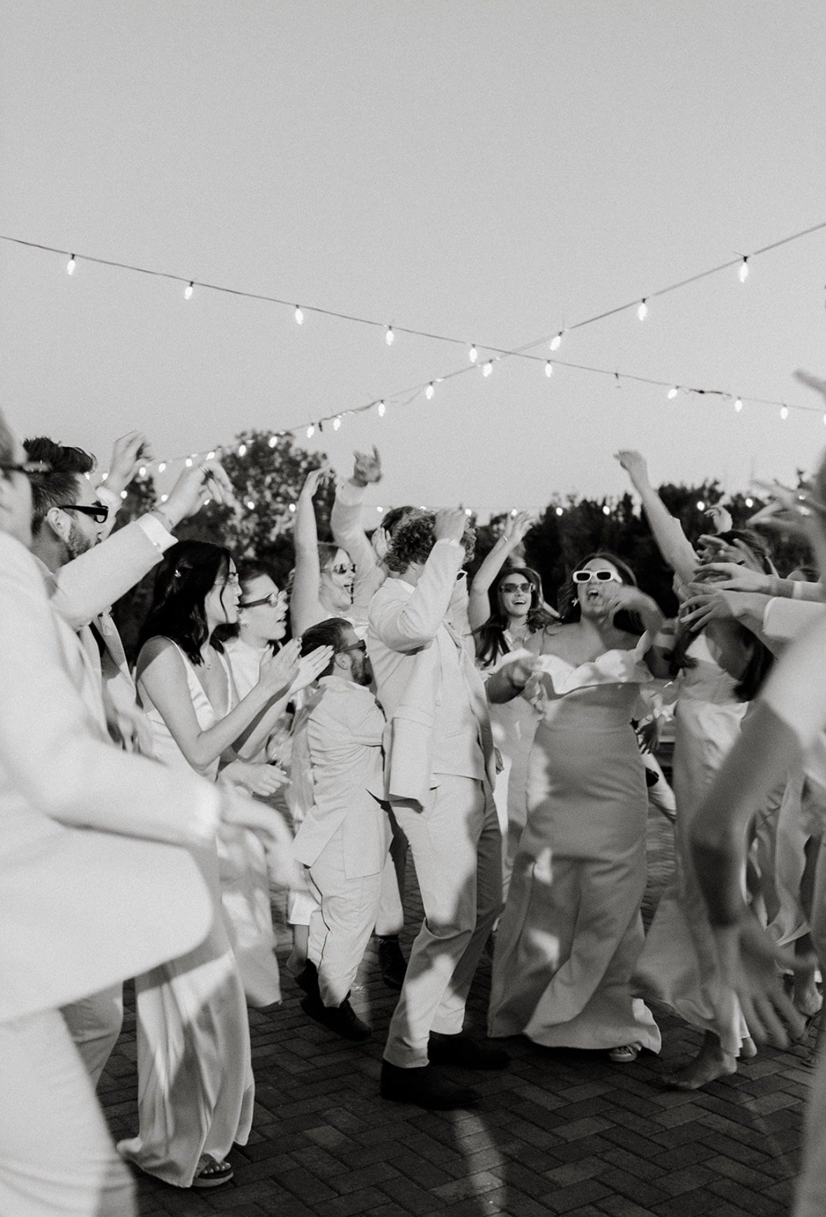 People dancing and celebrating outdoors at a wedding under string lights, with some wearing sunglasses.