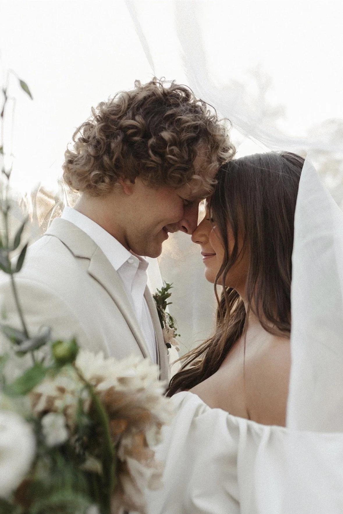 A close-up of a newlywed couple with their foreheads touching, smiling, under a white veil, with blurred greenery and flowers in the foreground.