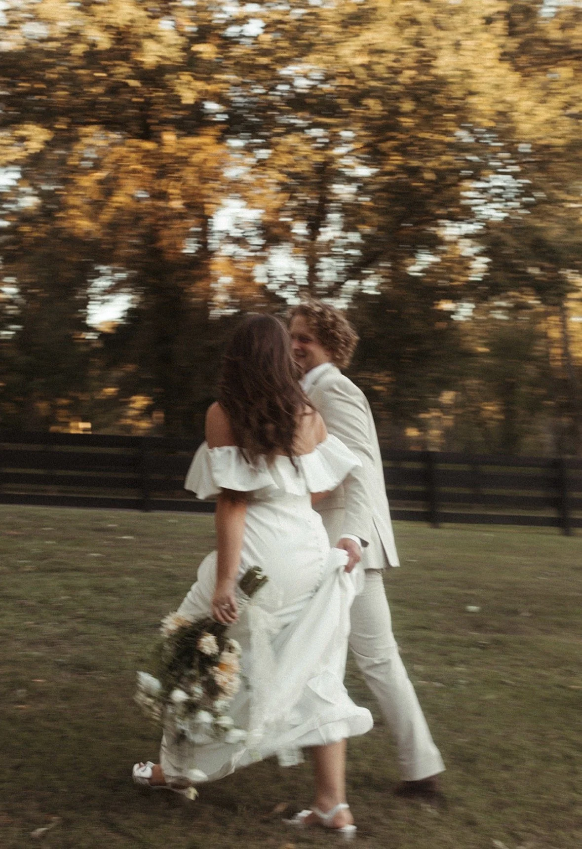 A couple in wedding attire dancing outdoors at sunset with autumn trees in the background.