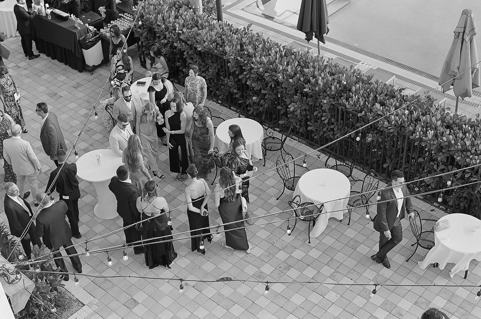 A balcony with people socializing, standing around round tables, some with drinks; string lights hang overhead, and there is a hedge and railing separating the balcony from the pool below.
