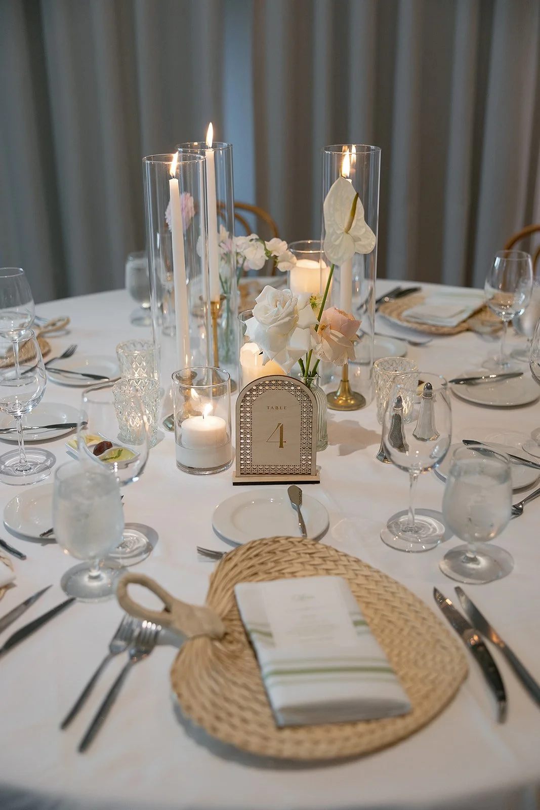 Elegant dining table decorated with white floral centerpieces, tall candle holders, and table number 4 sign, set with glassware, plates, and silverware for a formal event.
