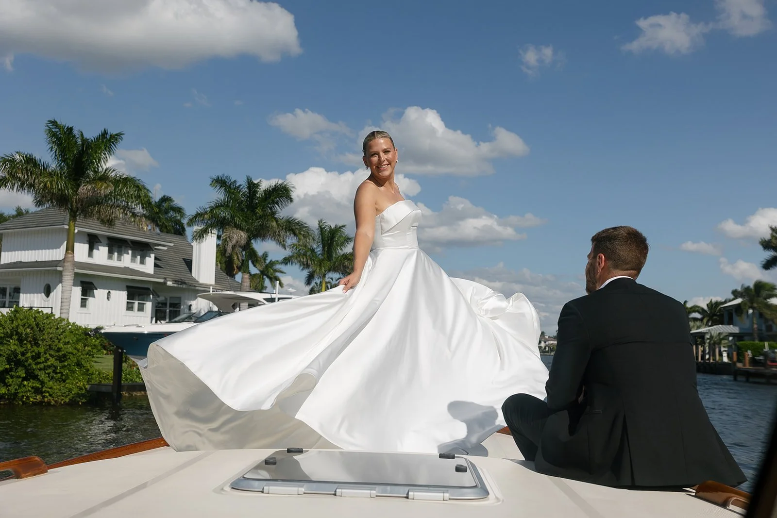 A bride in a white wedding dress is standing on a boat while a groom in a black suit kneels beside her on the boat, with palm trees, water, and houses in the background.