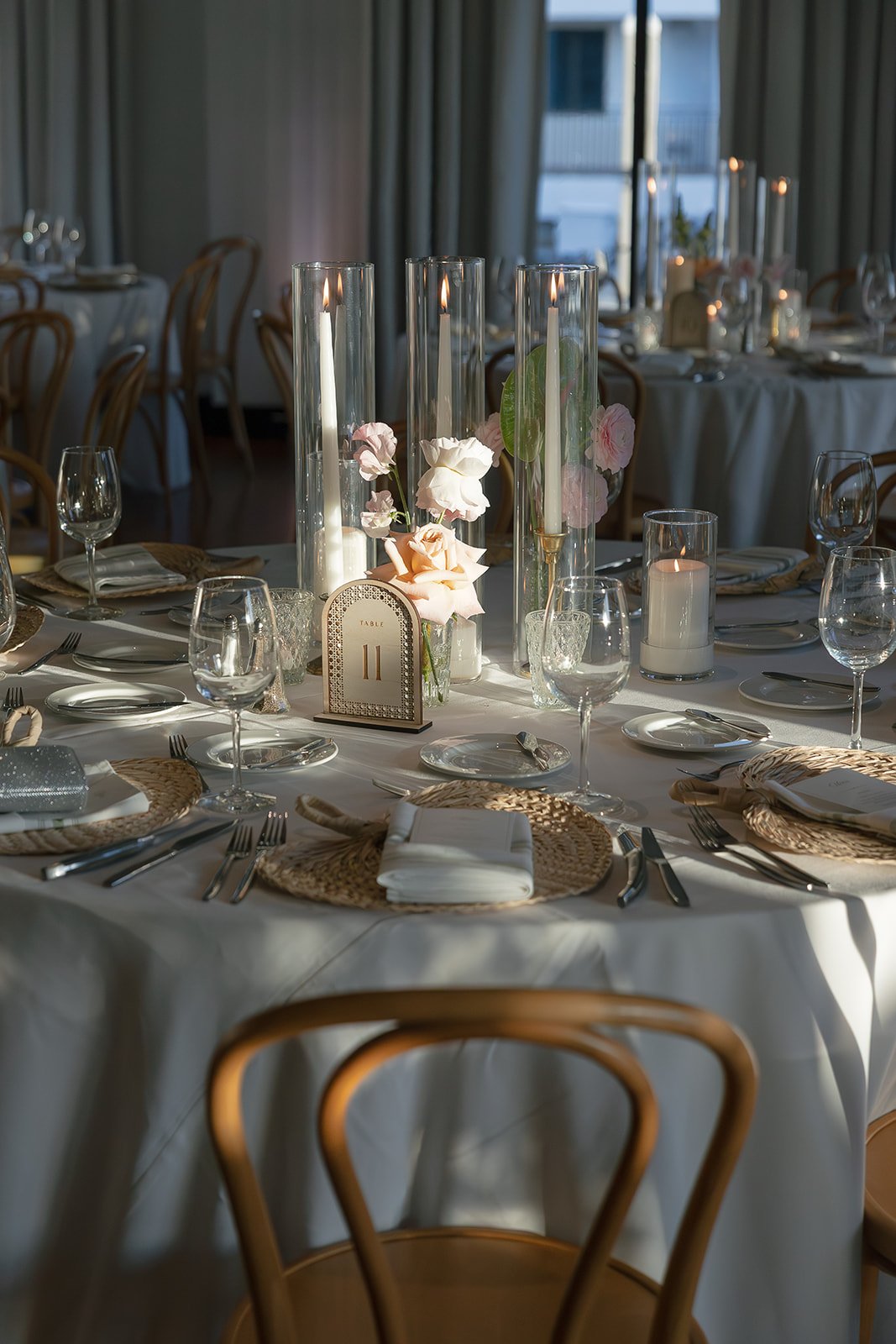Round banquet table draped with white tablecloth, set with woven placemats, white napkins, silverware, wine glasses, and candles, with a floral centerpiece and a table number sign labeled 'Table 11'.