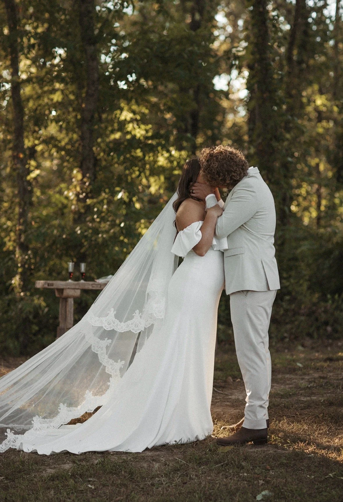 A bride and groom kiss outdoors during their wedding ceremony, with trees in the background and a small table with two glasses of wine in the distance.