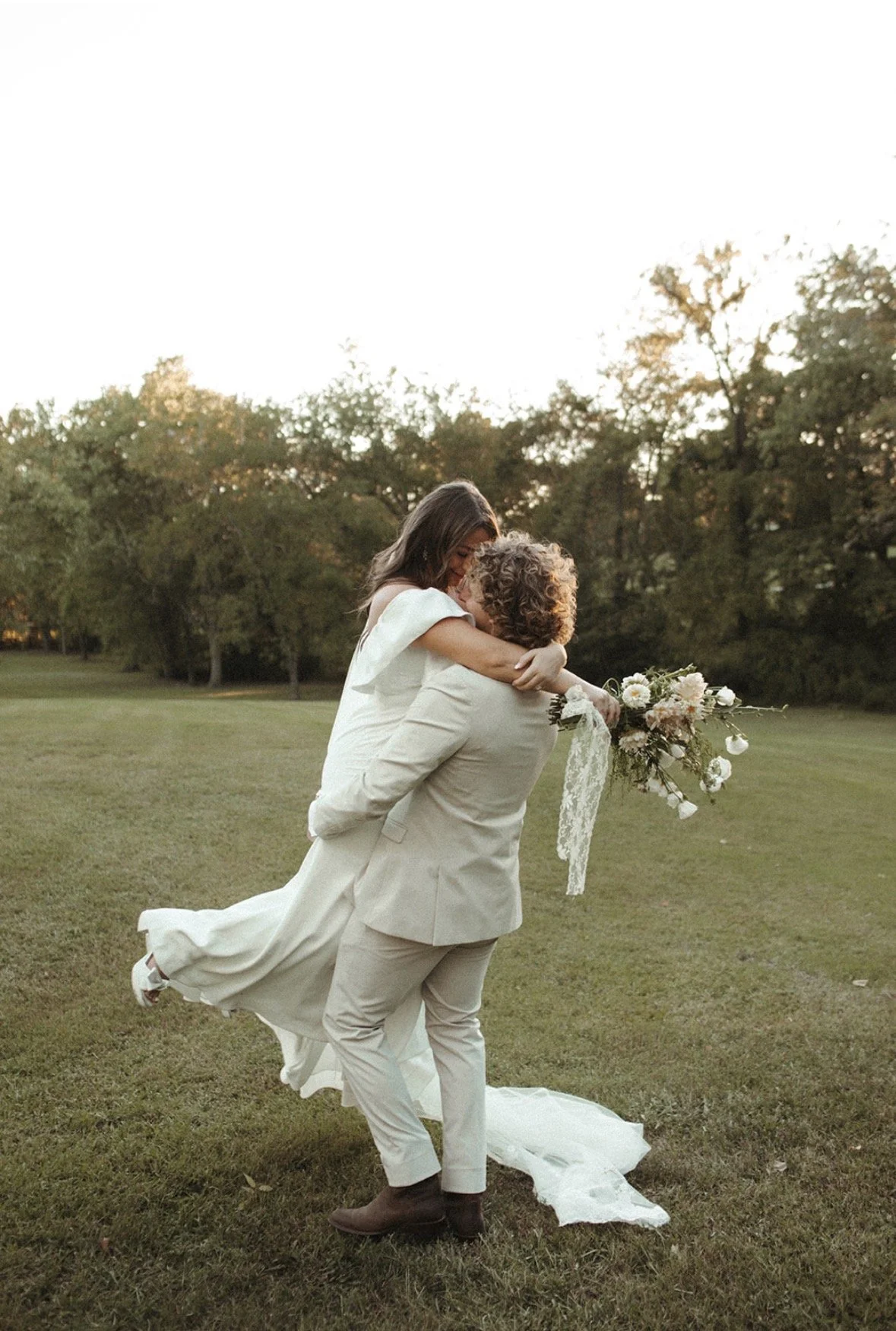 A man in a beige suit lifting a woman in a white wedding dress with a bouquet of white and pink flowers, outdoors on a grassy field with trees in the background.