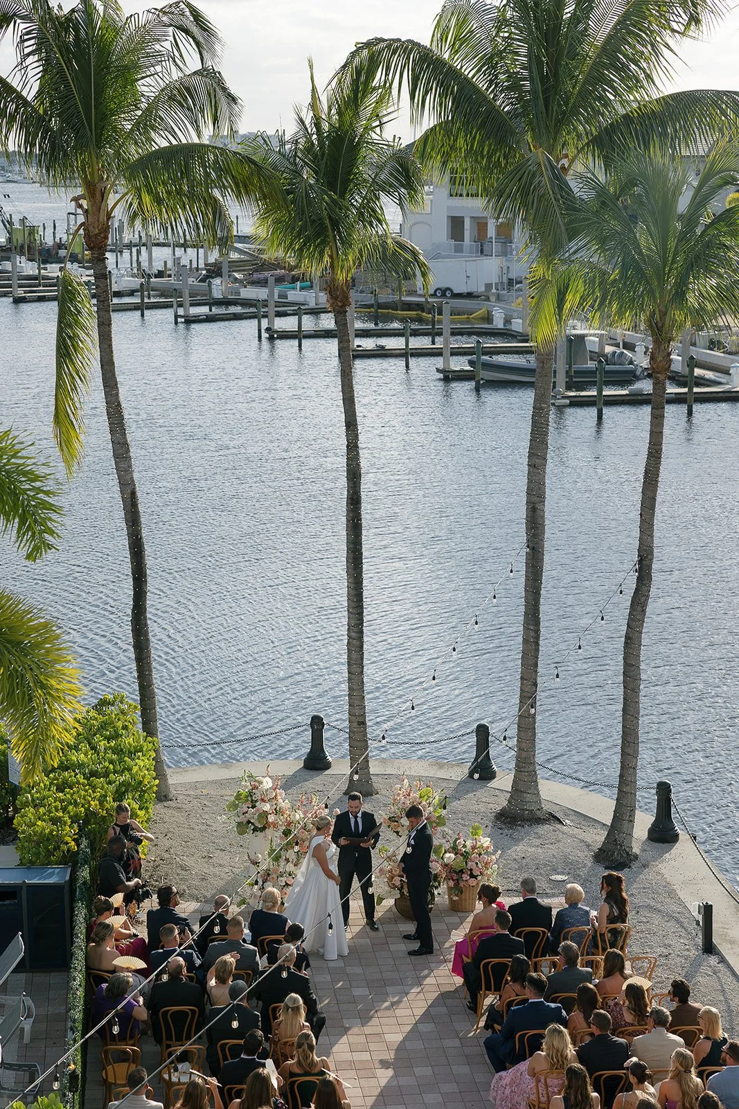 Outdoor wedding ceremony near a marina with palm trees and water, with guests seated in chairs and an officiant conducting the ceremony.