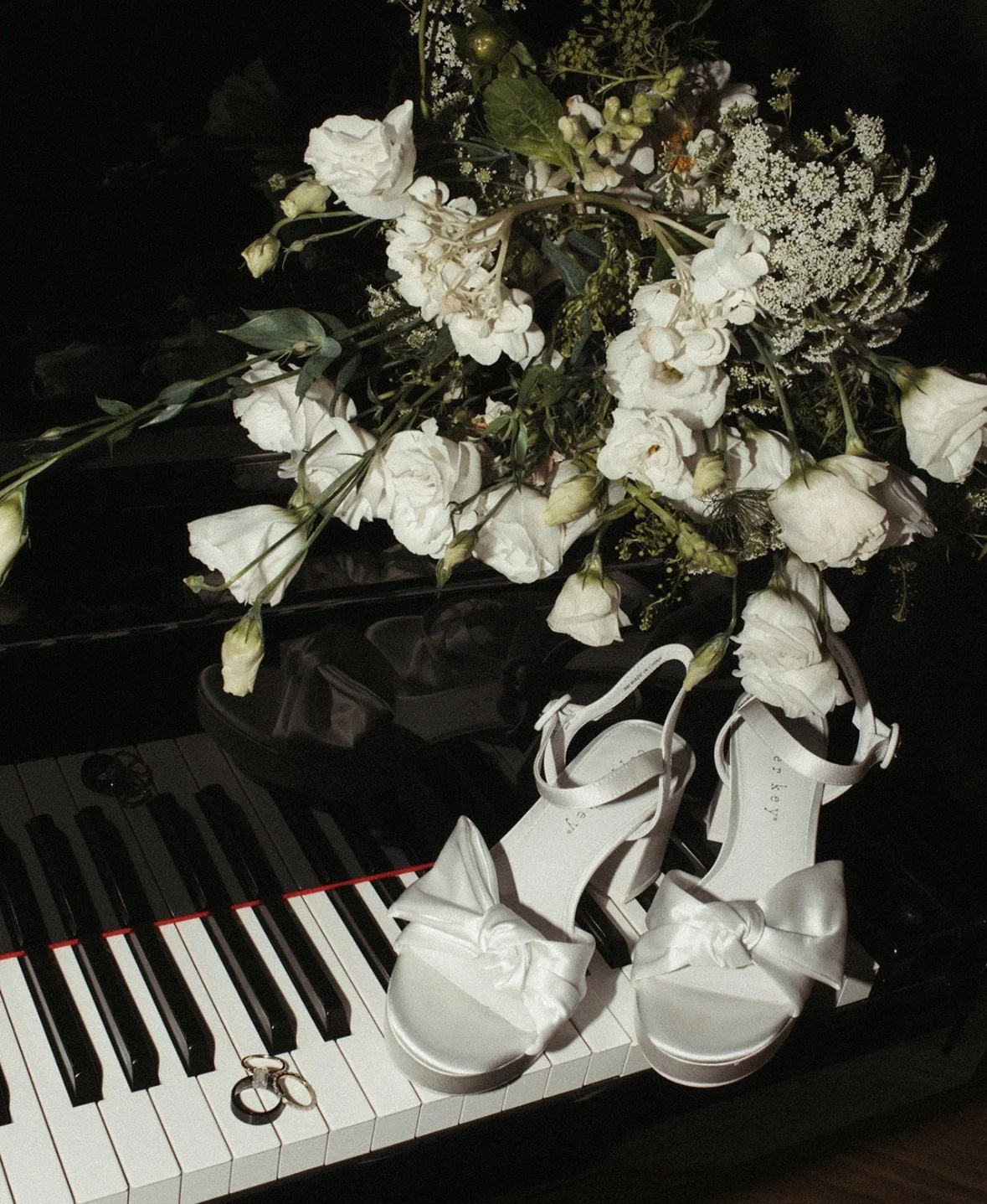White floral bouquet placed on a black piano with white high-heeled shoes featuring large bows on top of the keys, and two rings beside the shoes.