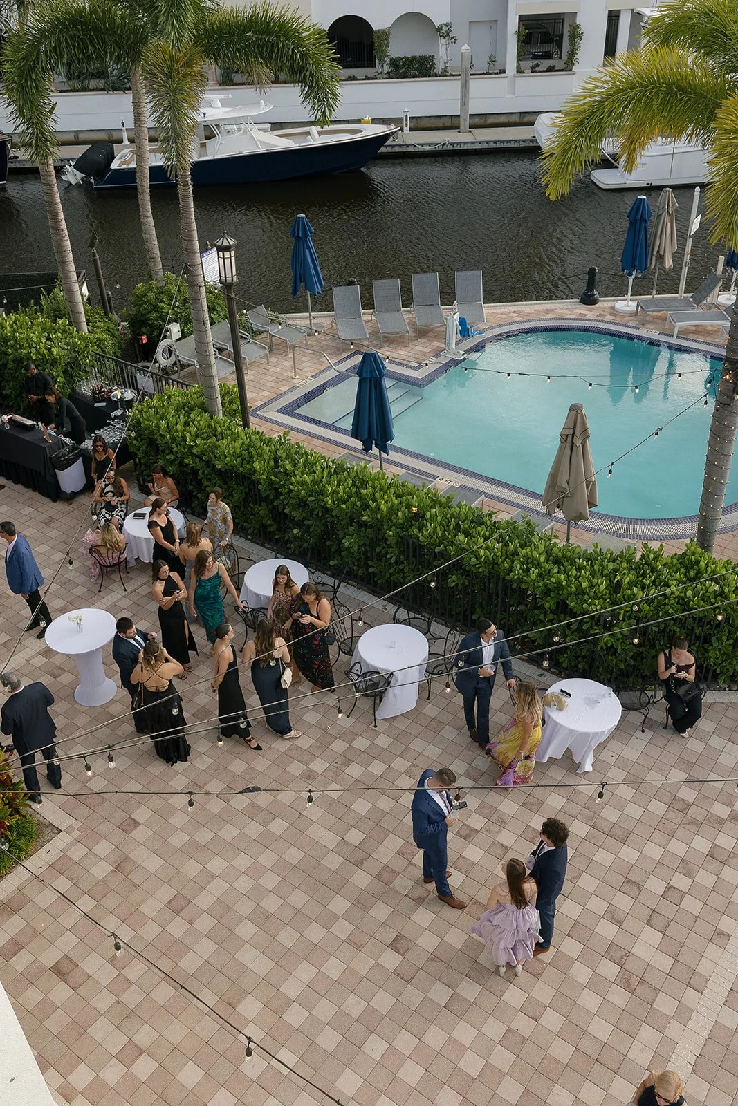 People gathered outdoors by a pool on a tiled patio, with some standing and chatting, others seated at tables with white tablecloths. There are lounge chairs, umbrellas, and greenery along the edge of the patio. A canal with boats is visible in the b